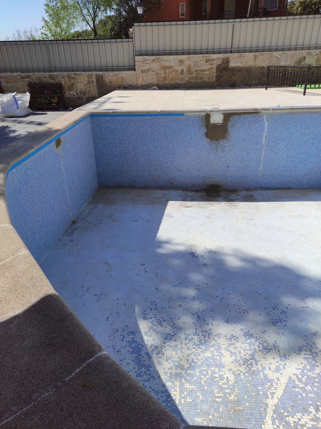 Piscina vacía, de azulejos de color azul claro, plataforma de hormigón, borde color tostado y una valla gris al fondo.