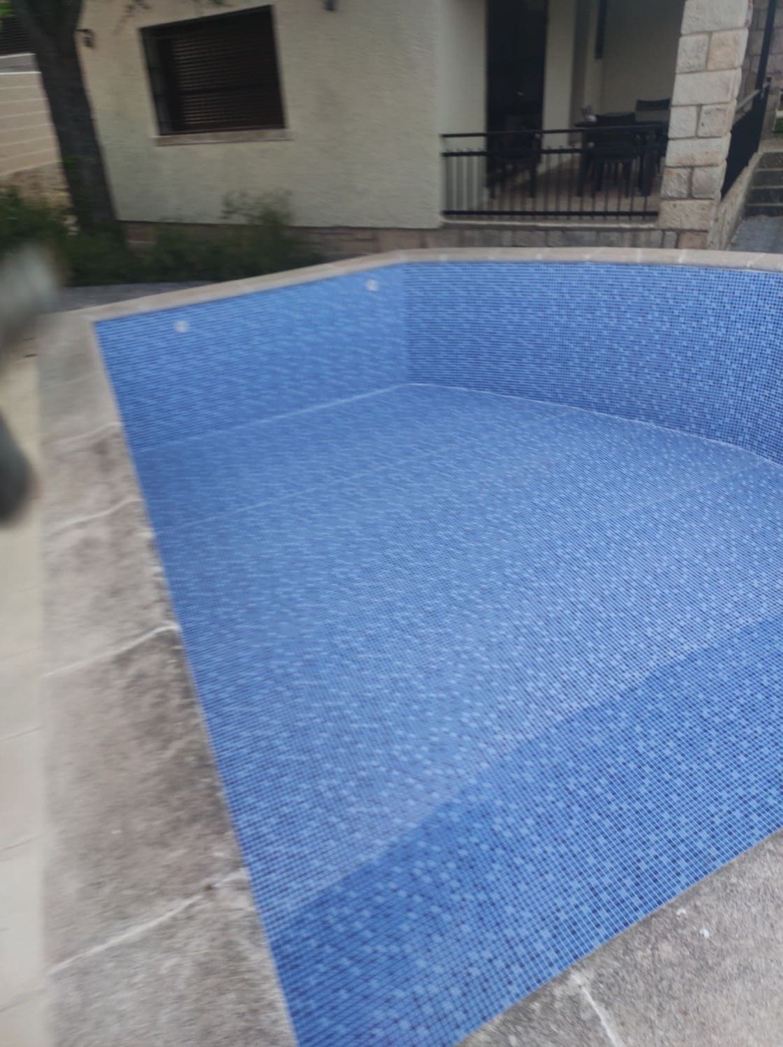 Piscina vacía de azulejos azules rodeada de un patio de piedra, al lado de un edificio.