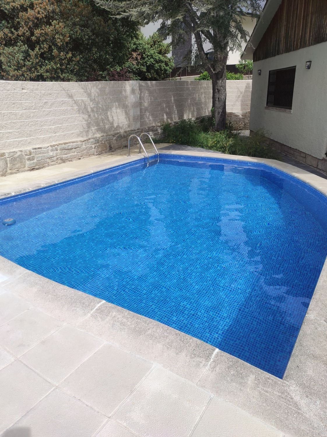 Piscina de azulejos azules rodeada de piedra gris. La piscina tiene una escalera; al fondo se ven un edificio y vegetación.