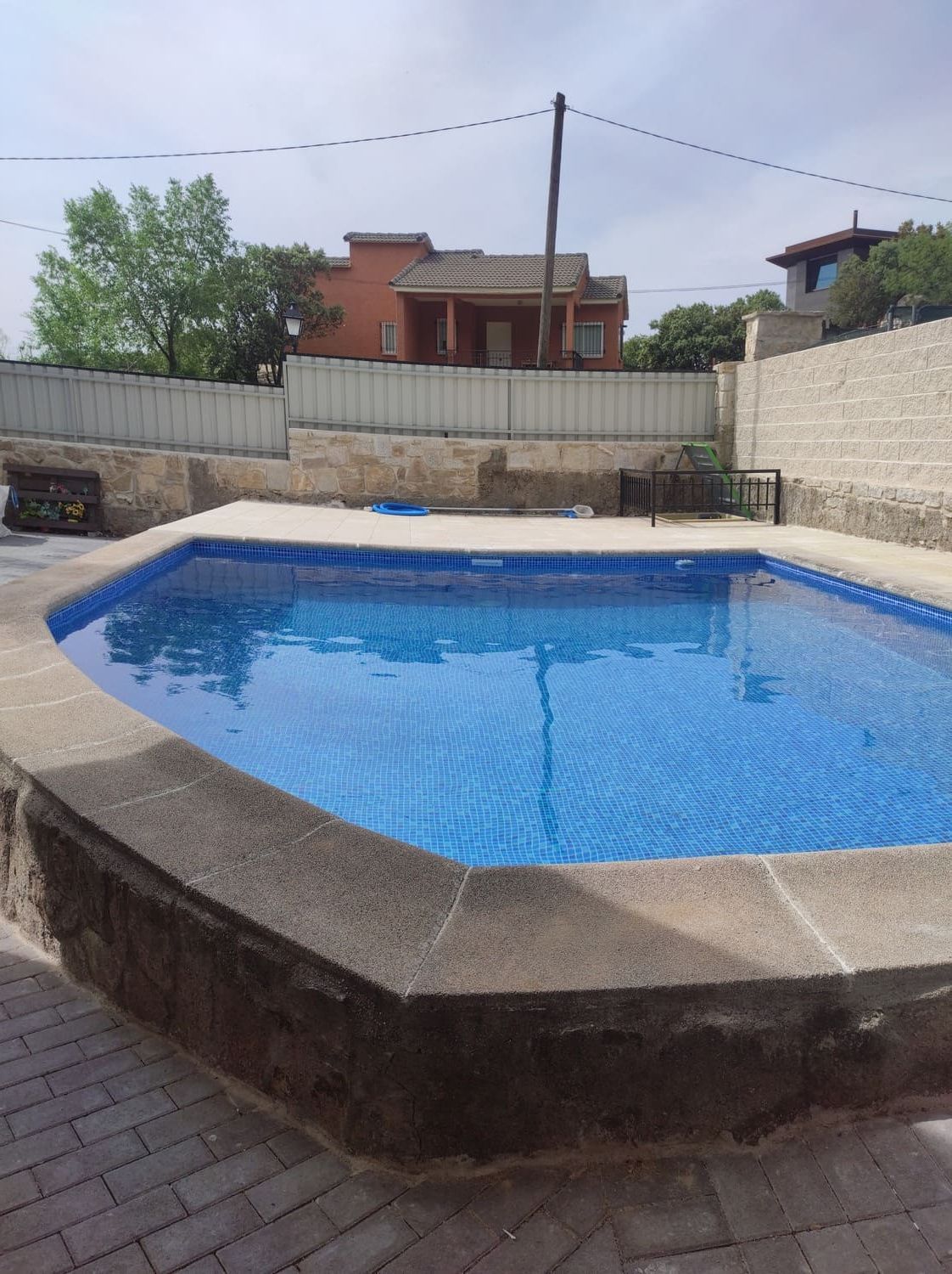 Una piscina rectangular de azulejos azules rodeada por un borde de piedra. Al fondo se ven edificios y el cielo.