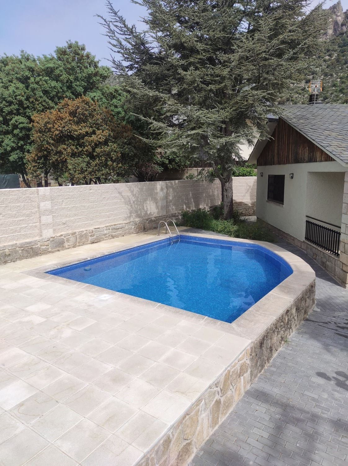 Piscina con agua azul y patio de piedra junto a una casa con techo de madera, árboles al fondo.