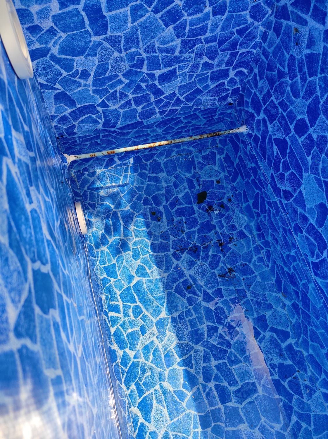 Interior de la piscina con mosaico azul. Se ven manchas oscuras en el fondo, con tubería y molduras blancas.