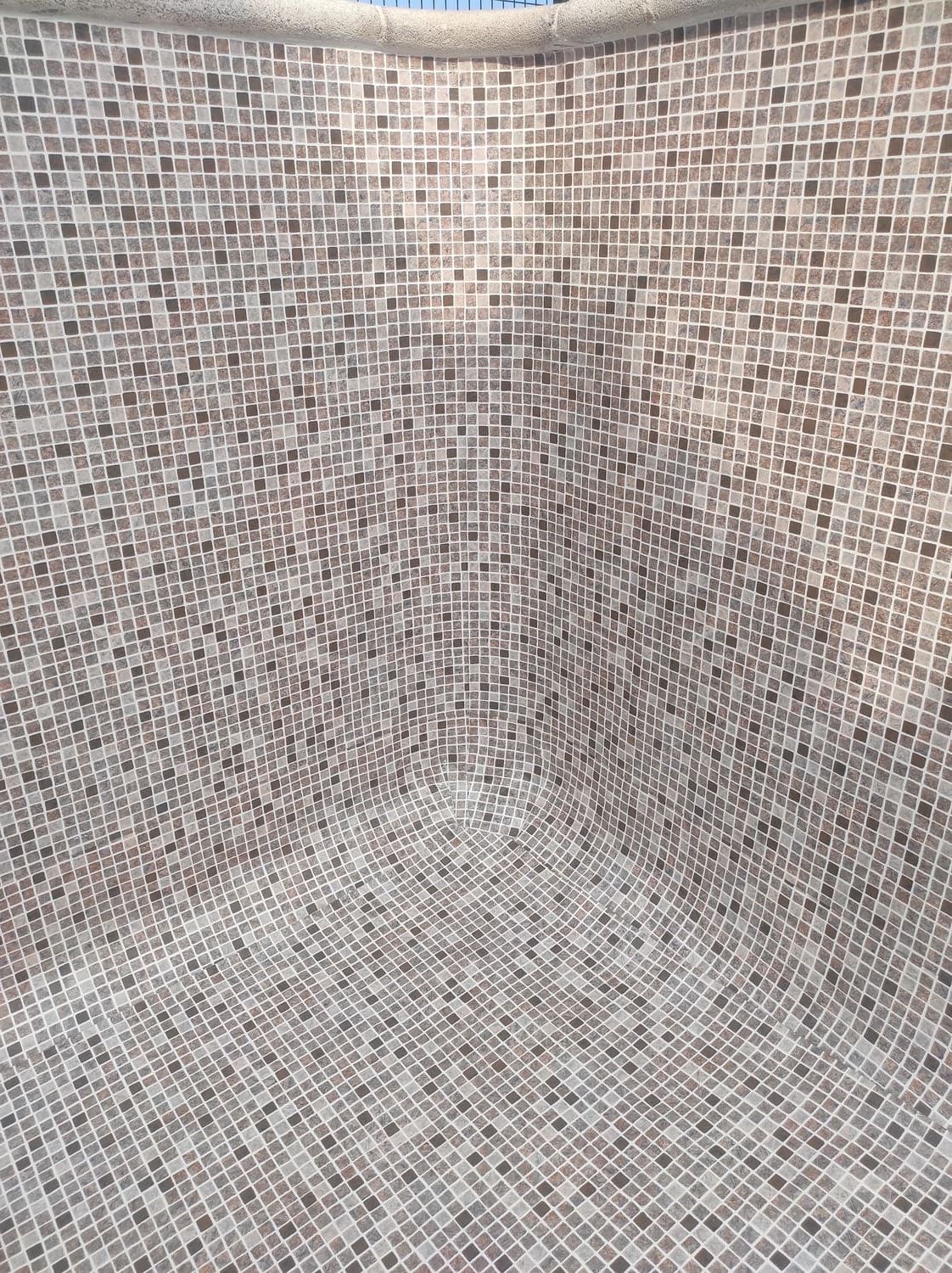 Esquina interior de una estructura de azulejos, probablemente una piscina o spa. Mosaicos beige y marrón cubren las paredes y el suelo.