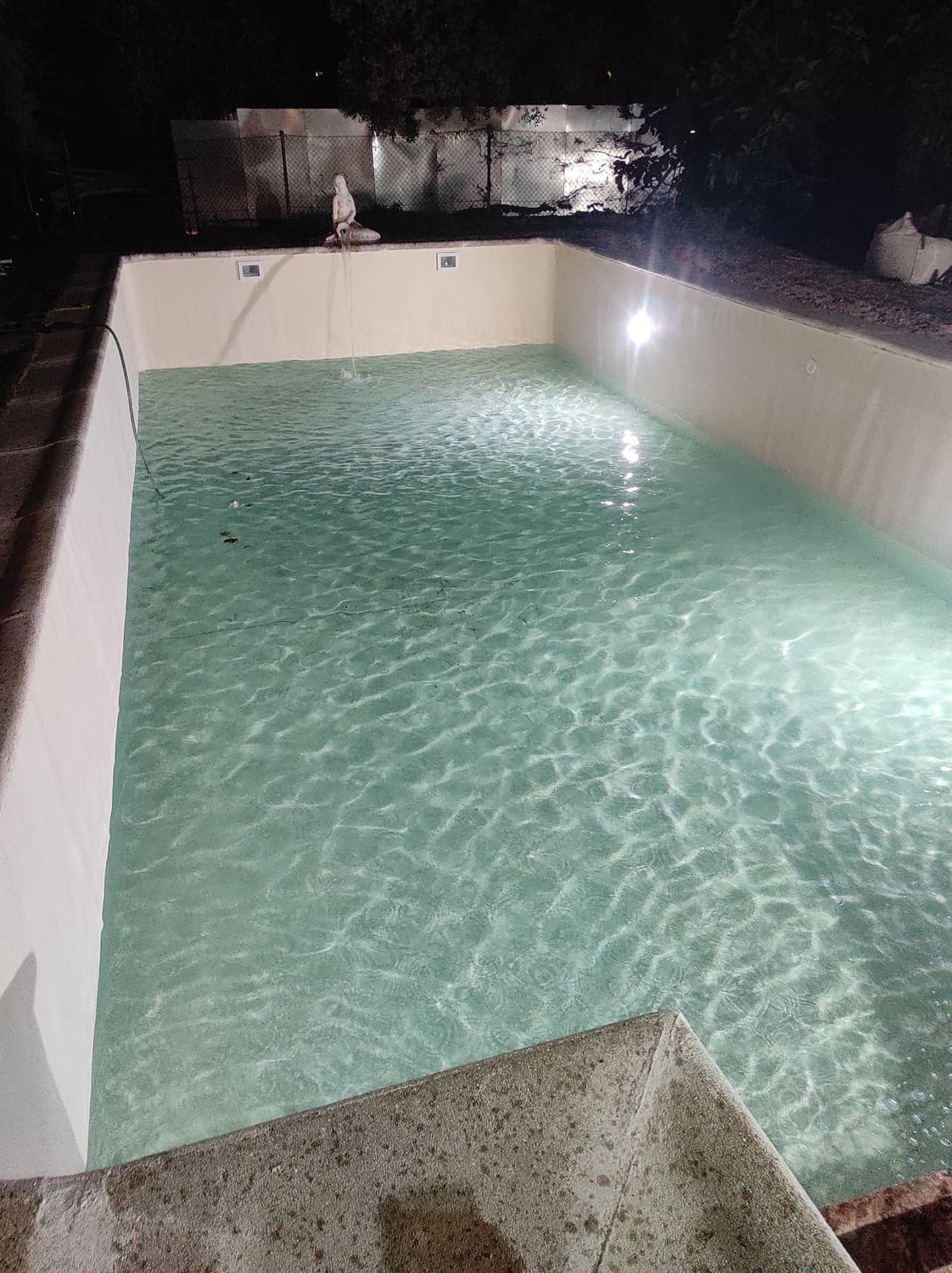 Piscina iluminada de noche. Paredes blancas, agua turquesa y escalones de piedra.