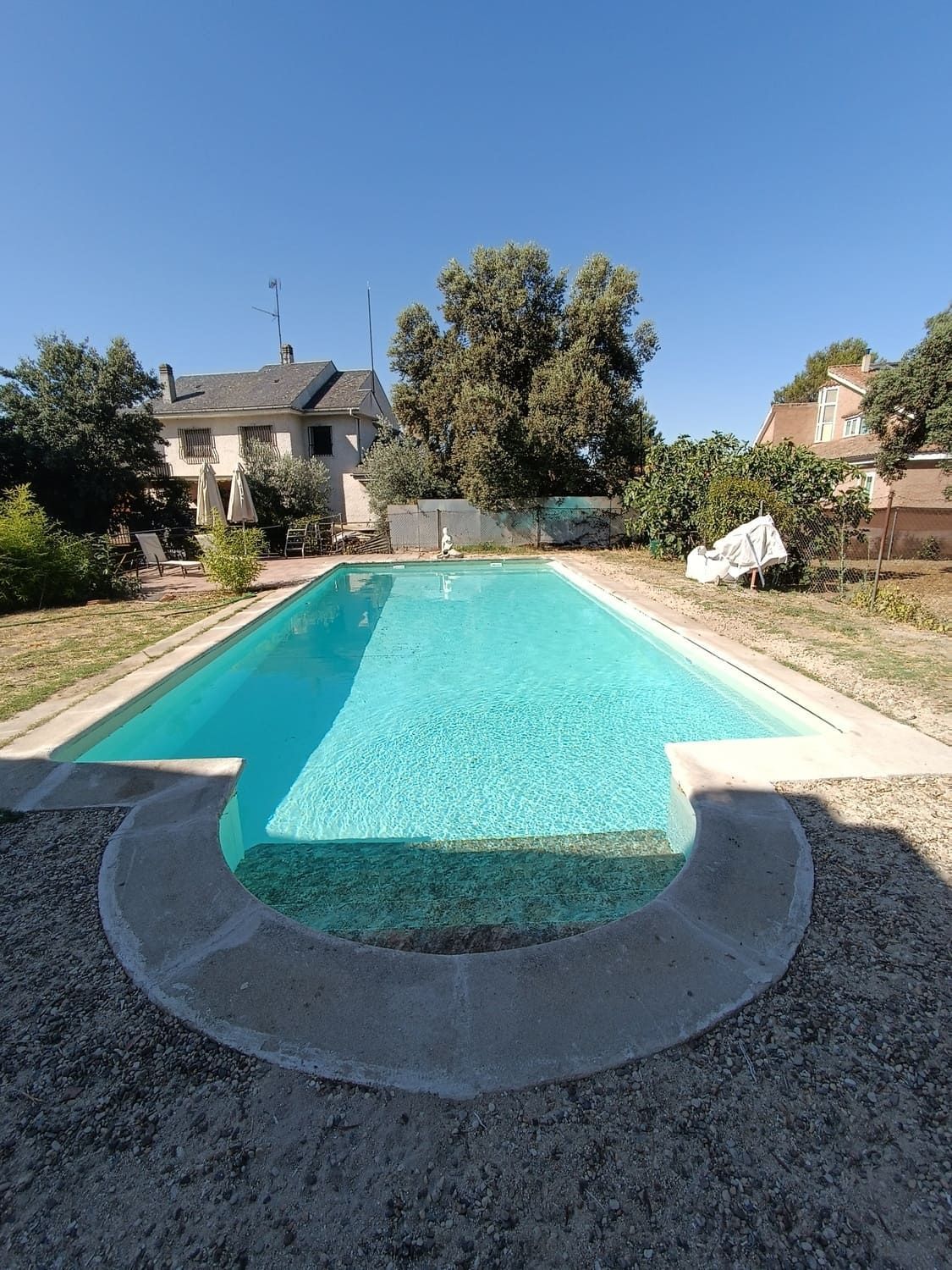 Piscina rectangular en el patio trasero, con casa al fondo. Agua azul, borde de hormigón, suelo de grava, día soleado.