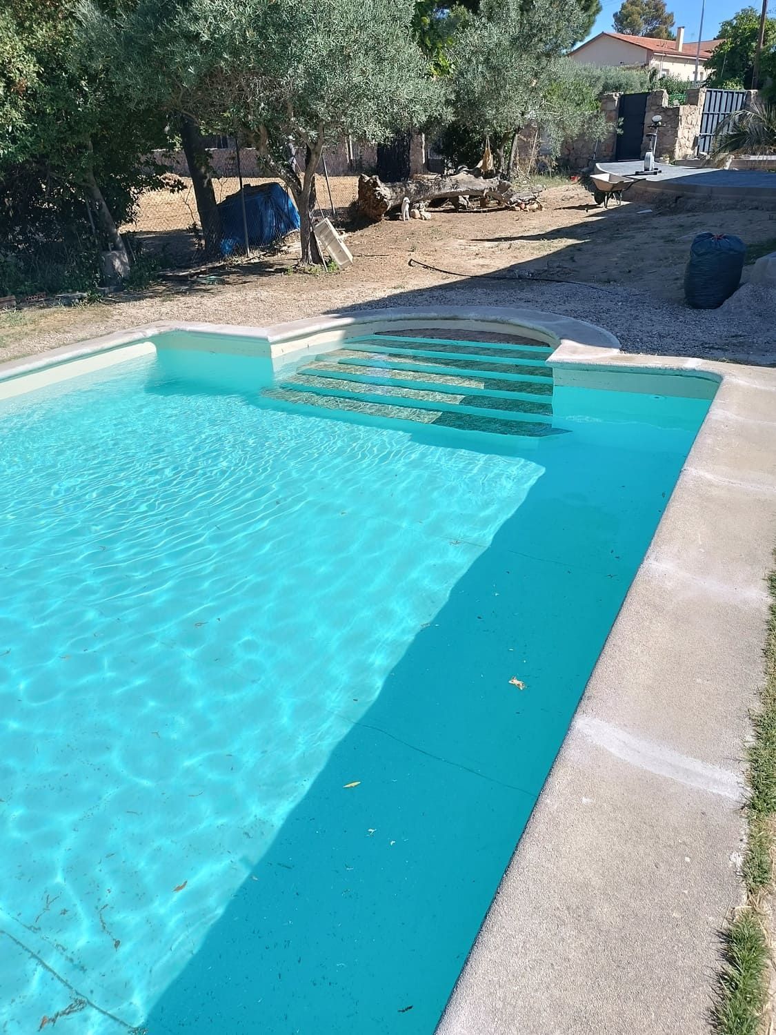 Una piscina rectangular con escalones incorporados, llena de agua turquesa clara, rodeada de hormigón y un patio.