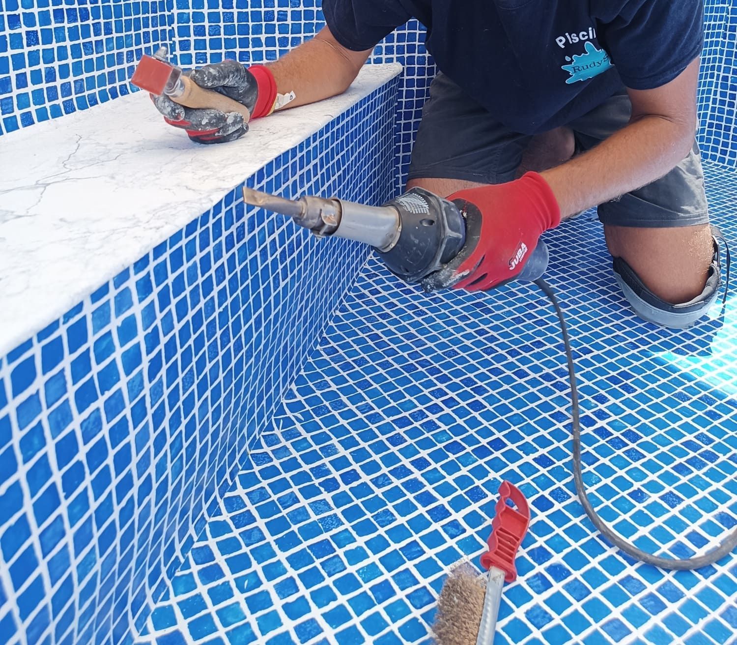 Una persona con guantes rojos utiliza una herramienta eléctrica en el borde de una piscina de azulejos.