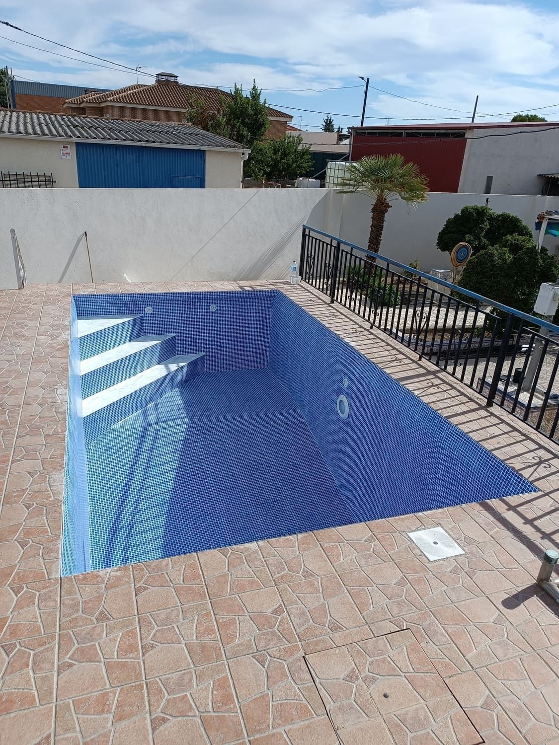 Piscina de azulejos azules con escalones, rodeada de pavimento de piedra, patio vallado y una pared blanca.