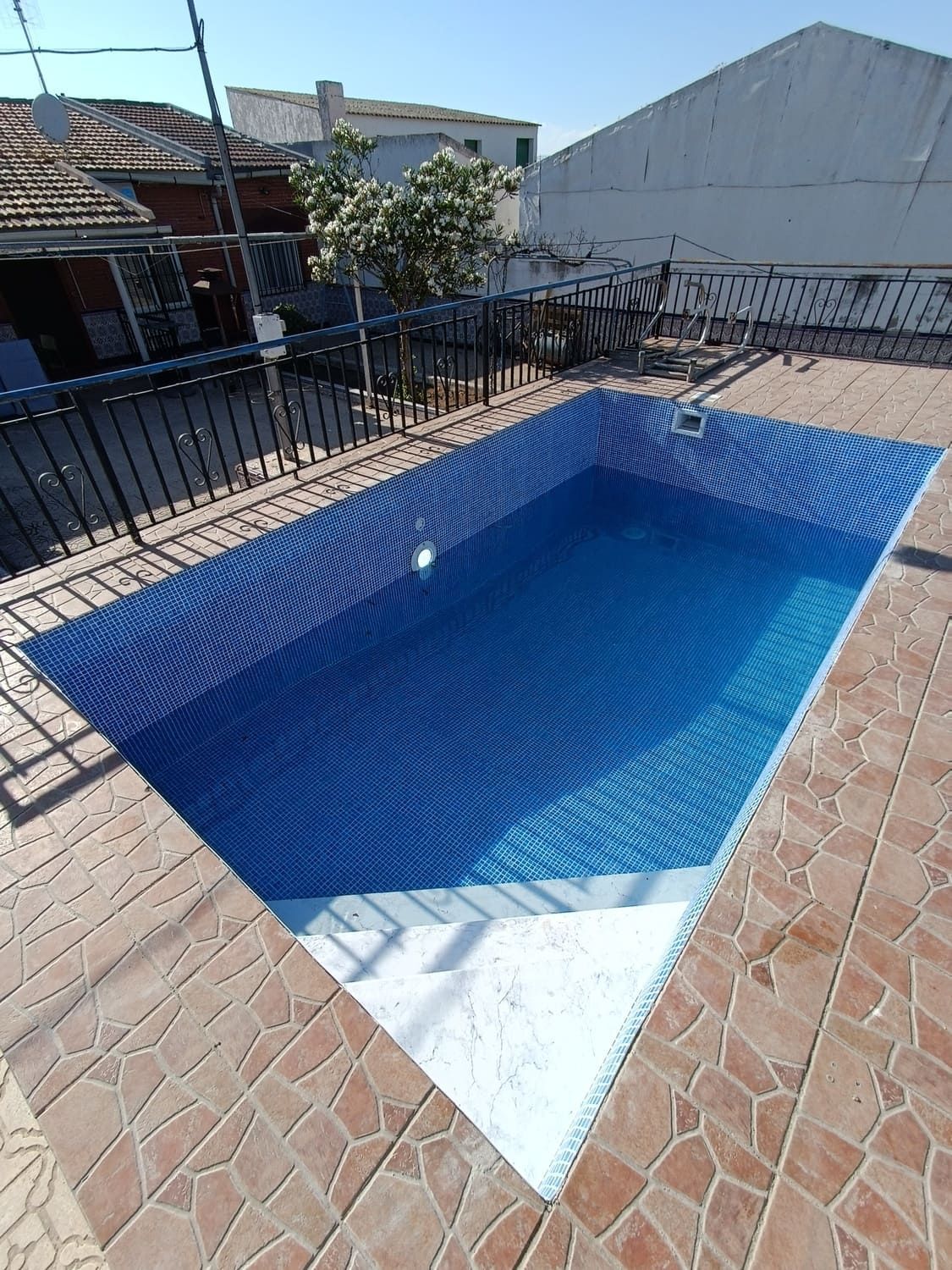 Piscina rectangular de azulejos azules con escalones blancos, rodeada por un patio de ladrillo y una valla negra.