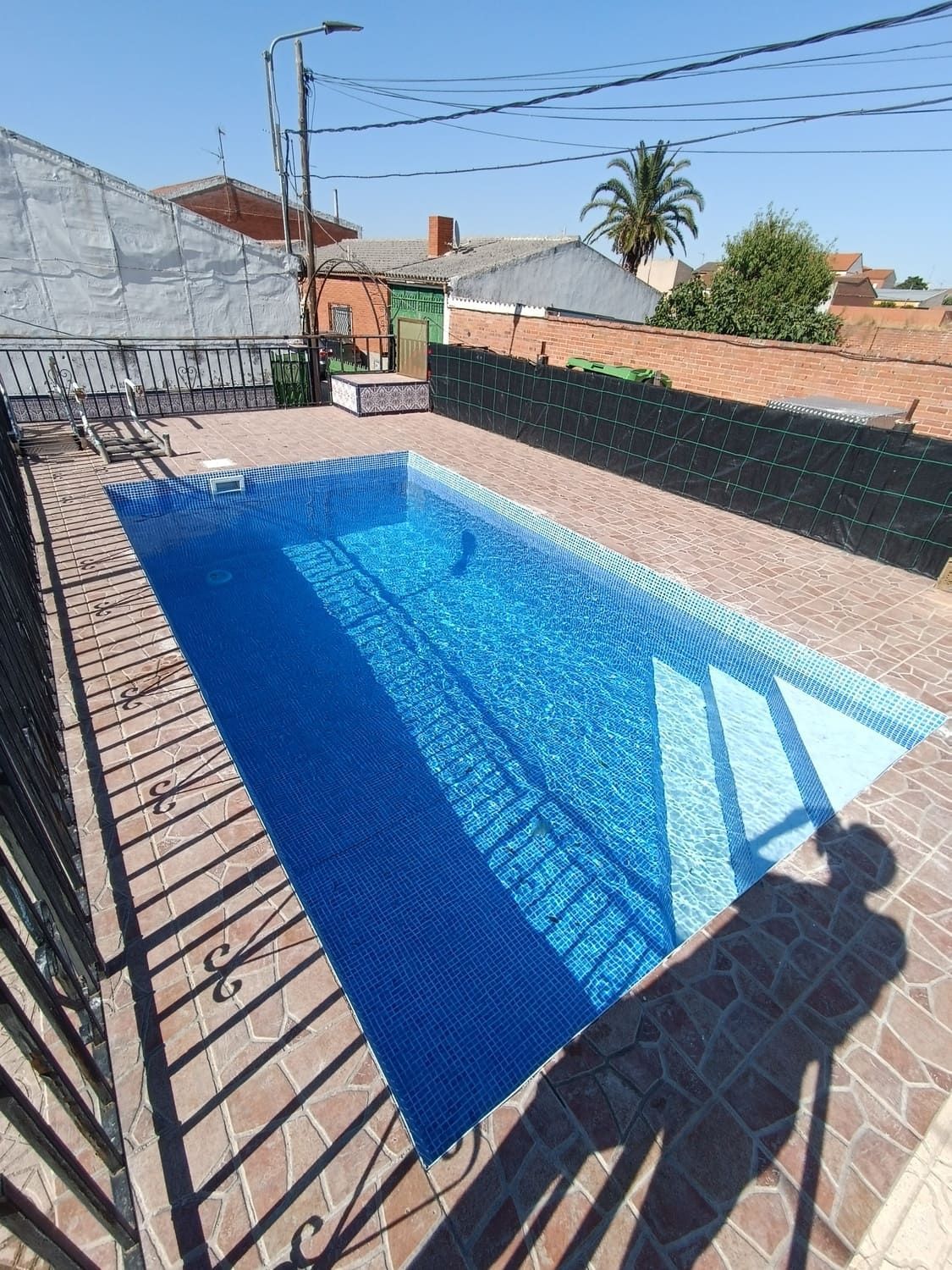 Piscina rectangular de azulejos azules rodeada de un patio de ladrillo. Al fondo, una valla negra y edificios, bajo un cielo radiante.