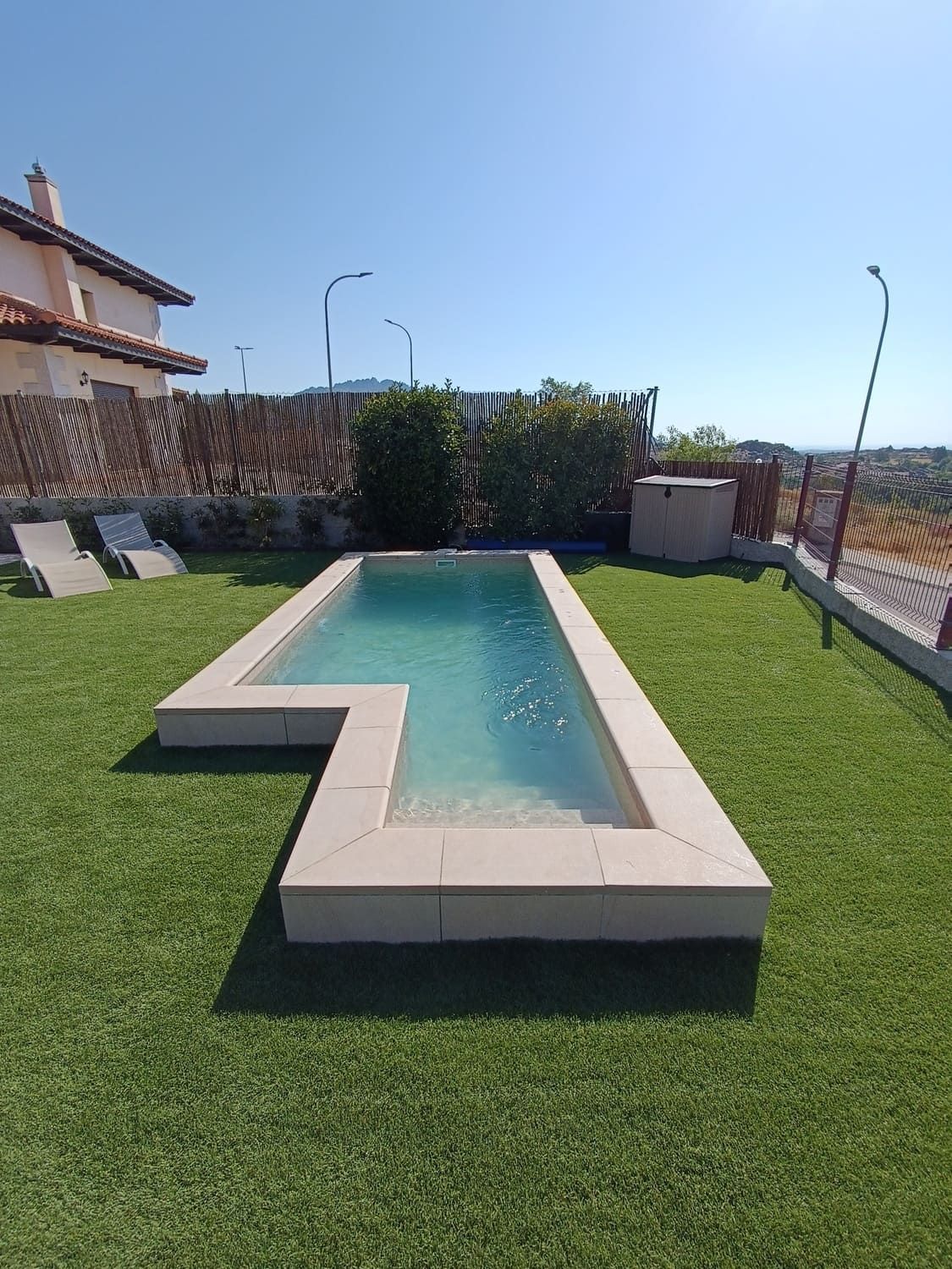 Piscina rectangular con agua azul claro y borde de piedra, sobre césped artificial verde. Casa al fondo.