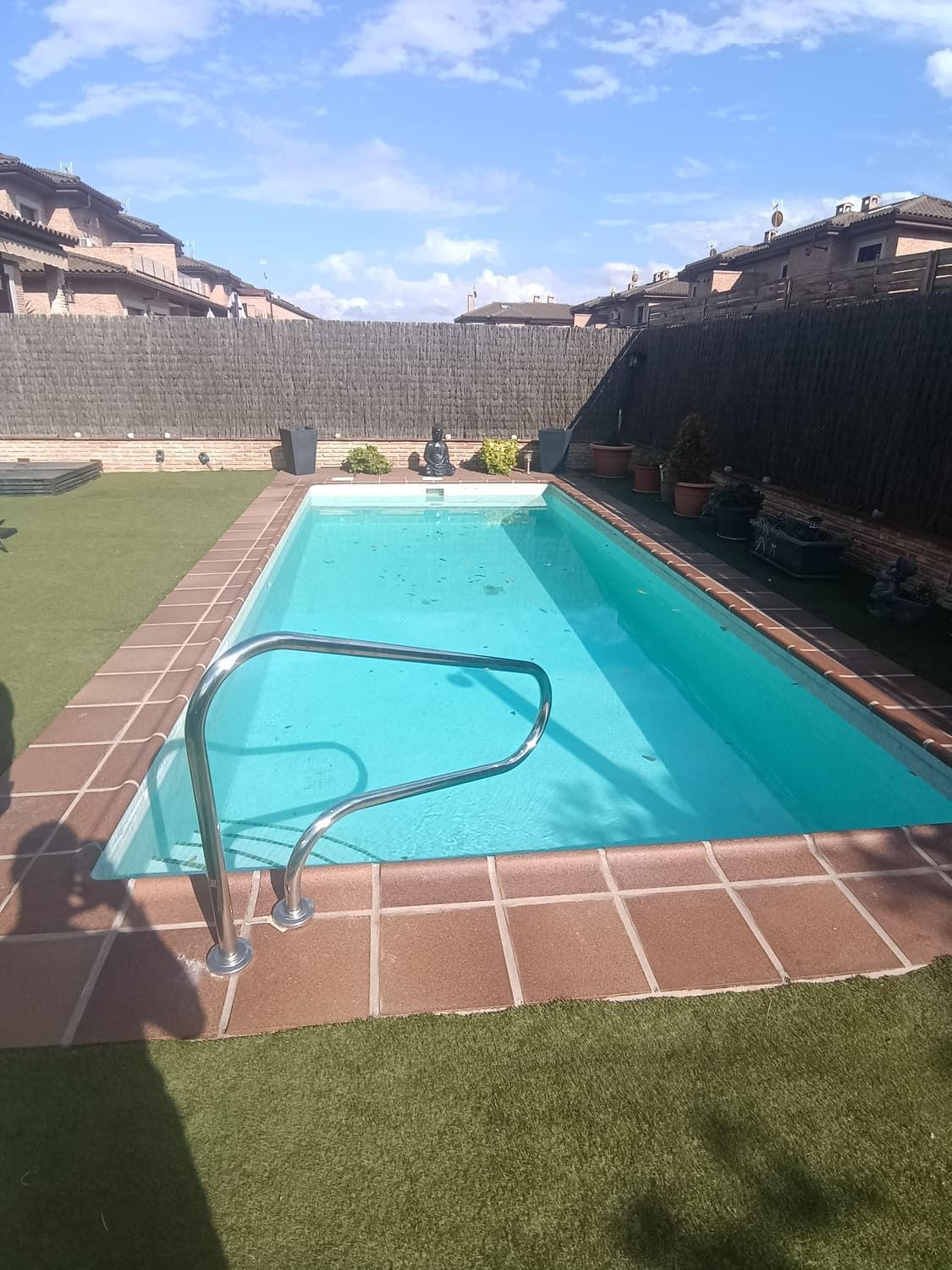 Piscina rectangular con barandilla metálica. Rodeada de baldosas marrones, césped y muro de piedra. Cielo azul y soleado.