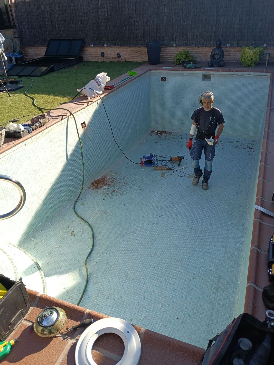 Hombre de pie en una piscina vacía, con herramientas esparcidas, en un patio trasero.