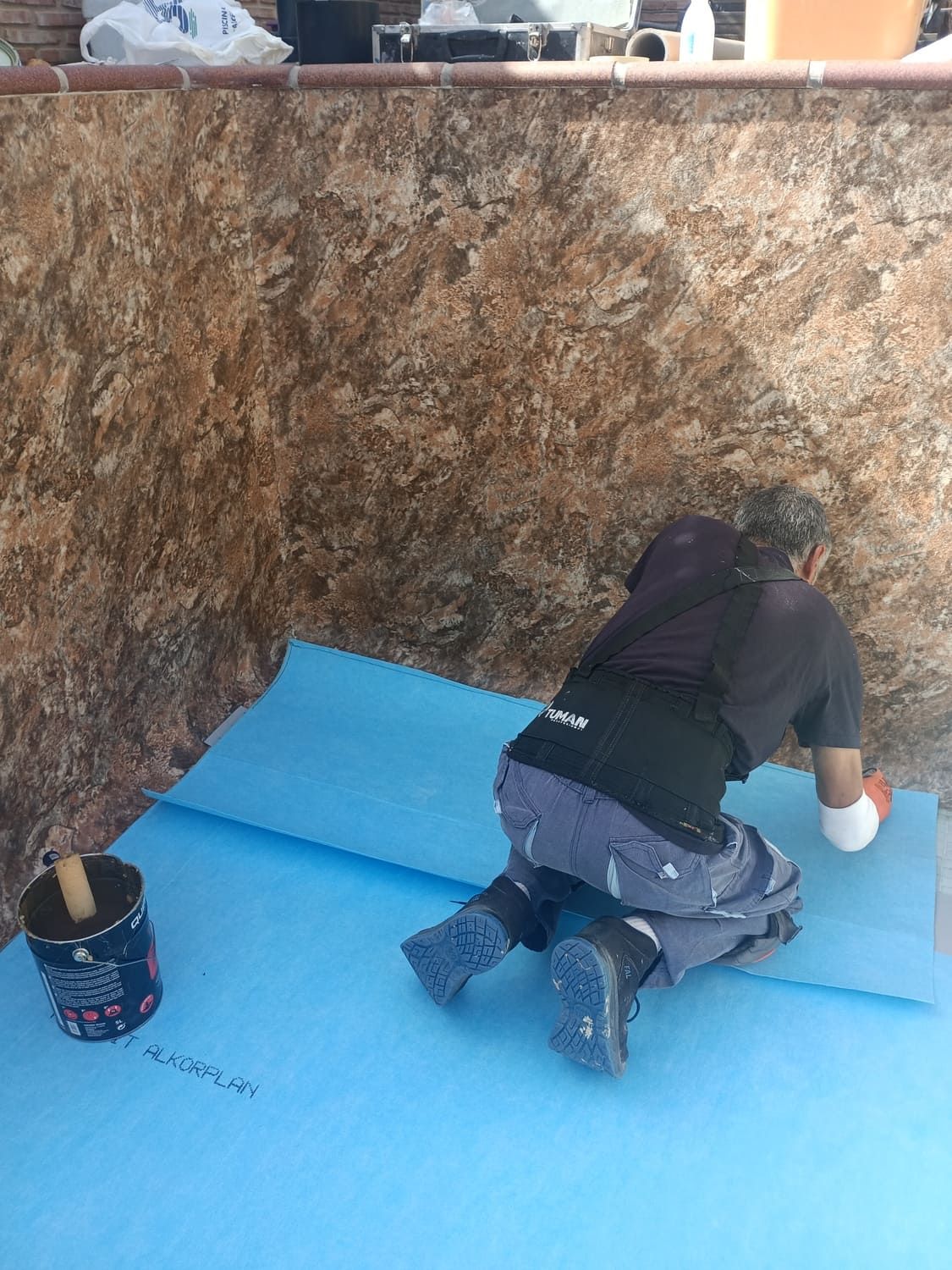 Persona arrodillada aplicando sellador a una lámina azul sobre un muro de piedra marrón. Cerca hay un cubo de sellador.