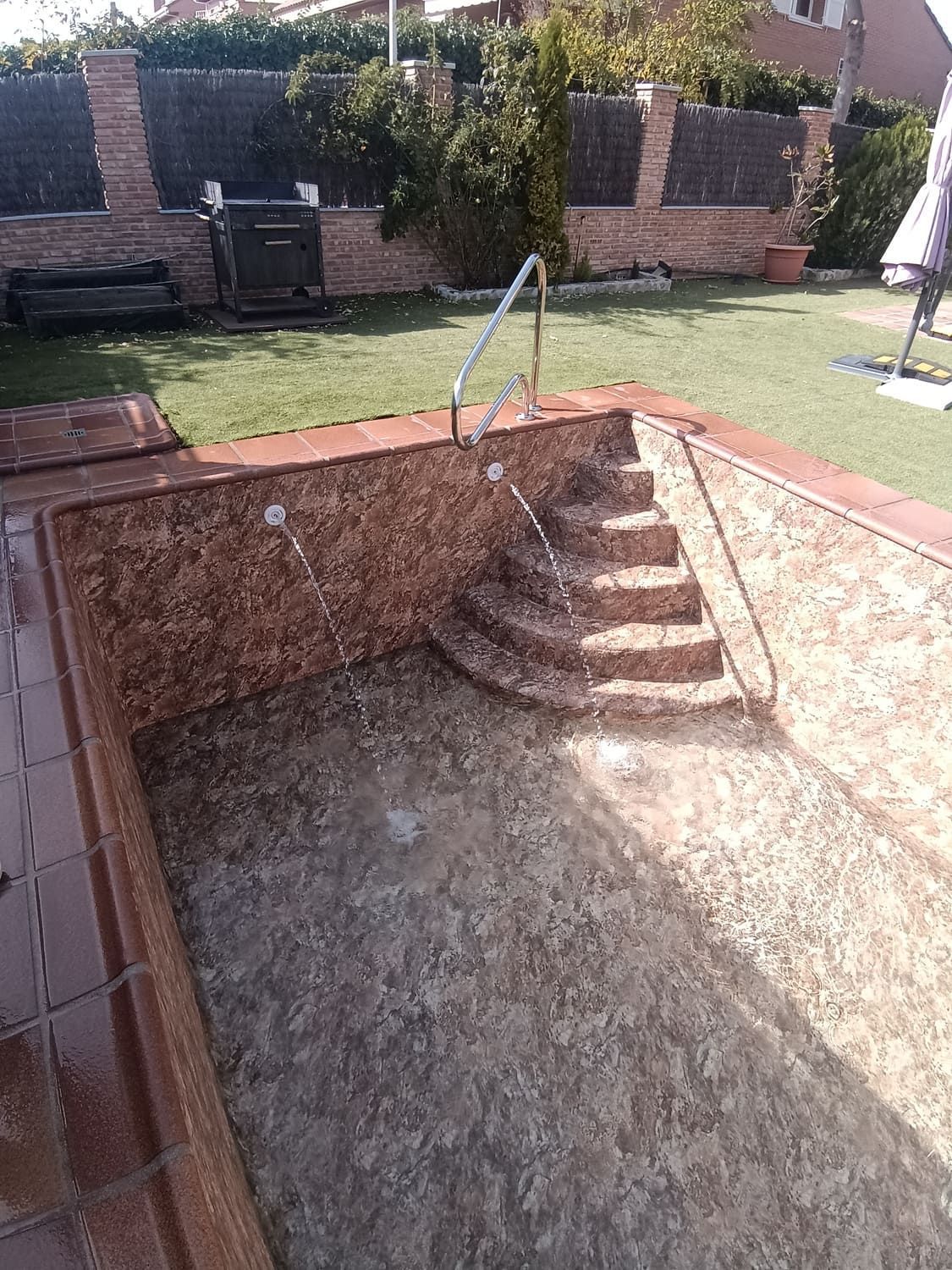 Piscina vacía con borde de azulejos marrones, escalones y pasamanos metálicos. Césped verde y pared de ladrillo al fondo.
