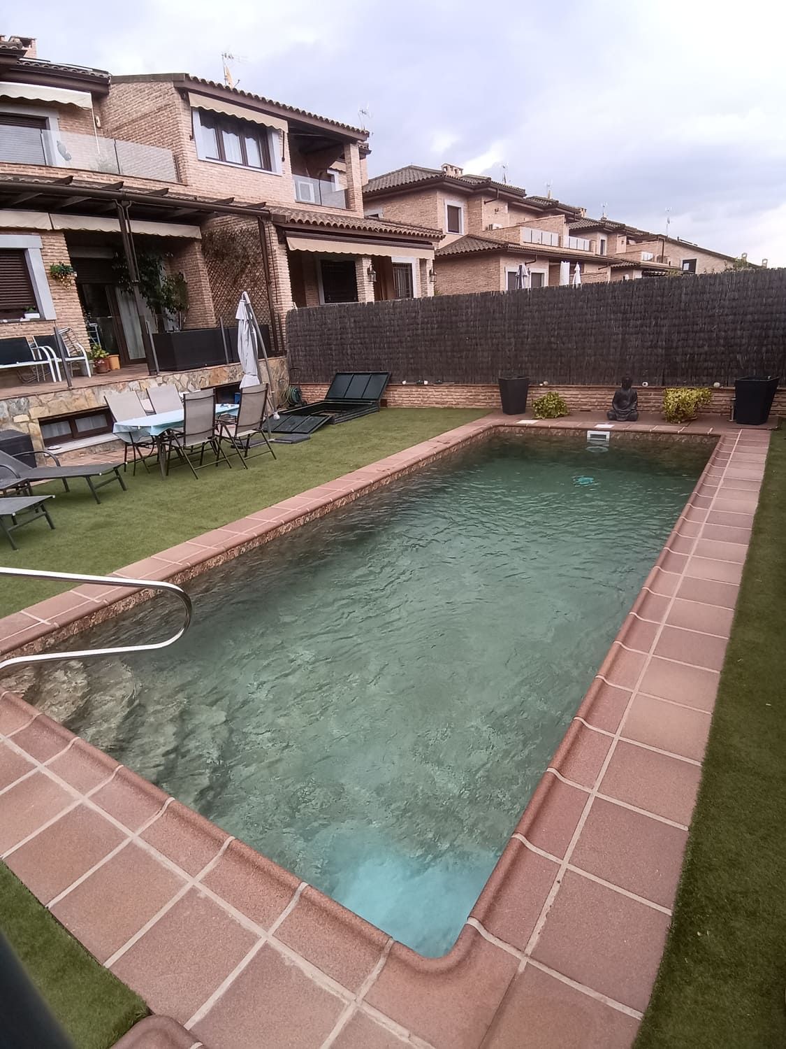 Una piscina rectangular con borde de ladrillo, frente a una hilera de casas adosadas. Césped verde la rodea.
