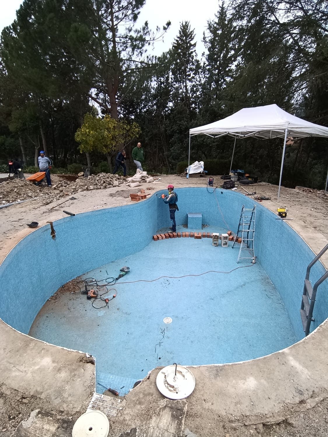 Piscina en construcción, revestida con mosaico azul. Trabajadores, carpa blanca y árboles al fondo.