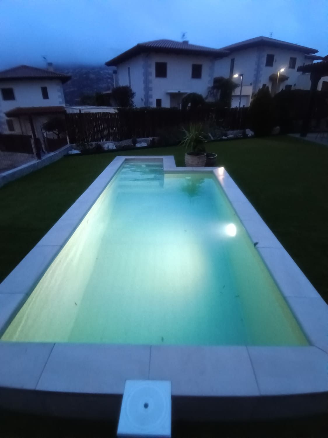 Una piscina rectangular iluminada al anochecer, rodeada de piedra clara. Casas y vegetación al fondo.