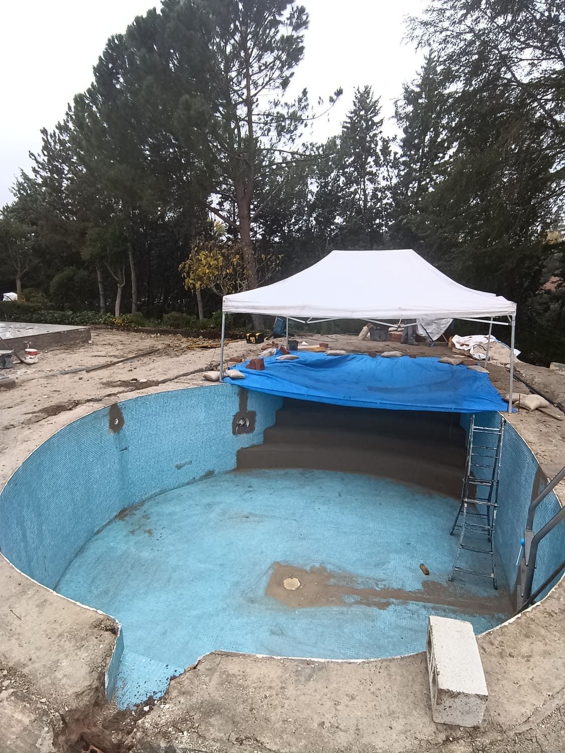 Una piscina ovalada, vacía y con líneas azules en construcción. Una carpa blanca se encuentra detrás de la piscina, con árboles al fondo.