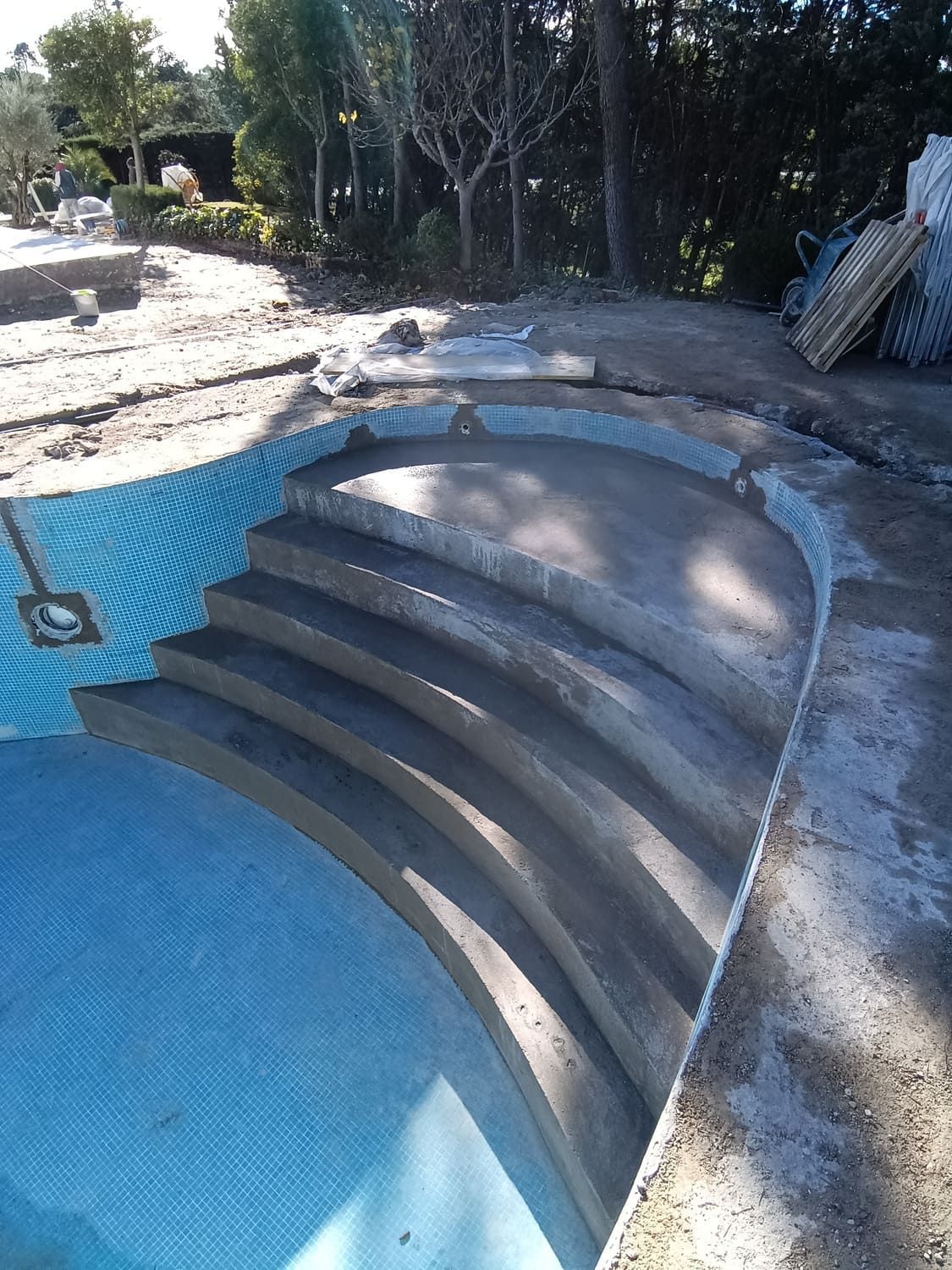 Escaleras de hormigón que conducen a una piscina parcialmente embaldosada. Hay materiales de construcción cerca.