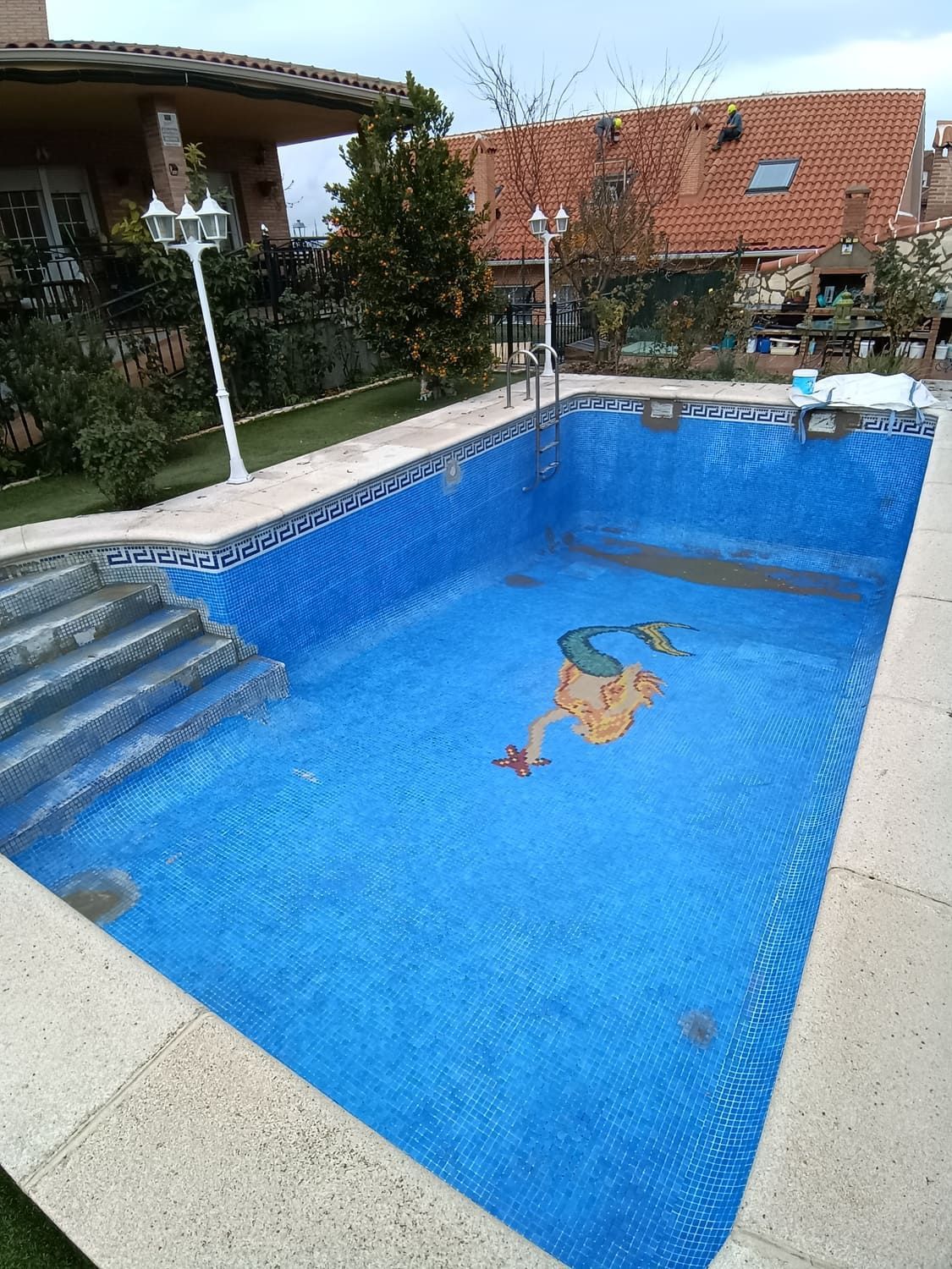 Piscina vacía de azulejos azules con mosaicos en el fondo, rodeada de hormigón beige y vegetación.