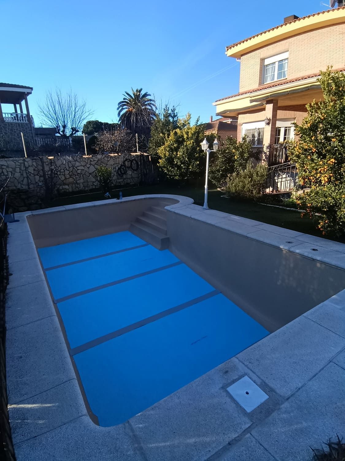 Piscina exterior vacía cubierta con lona azul, al lado de una casa de dos pisos, día soleado.