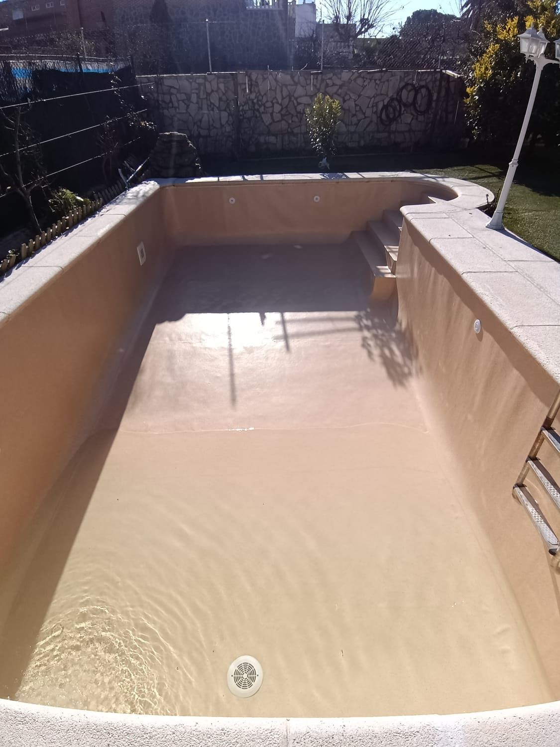 Piscina vacía, rectangular, de color marrón claro, con escalones, rodeada de piedra y césped verde.