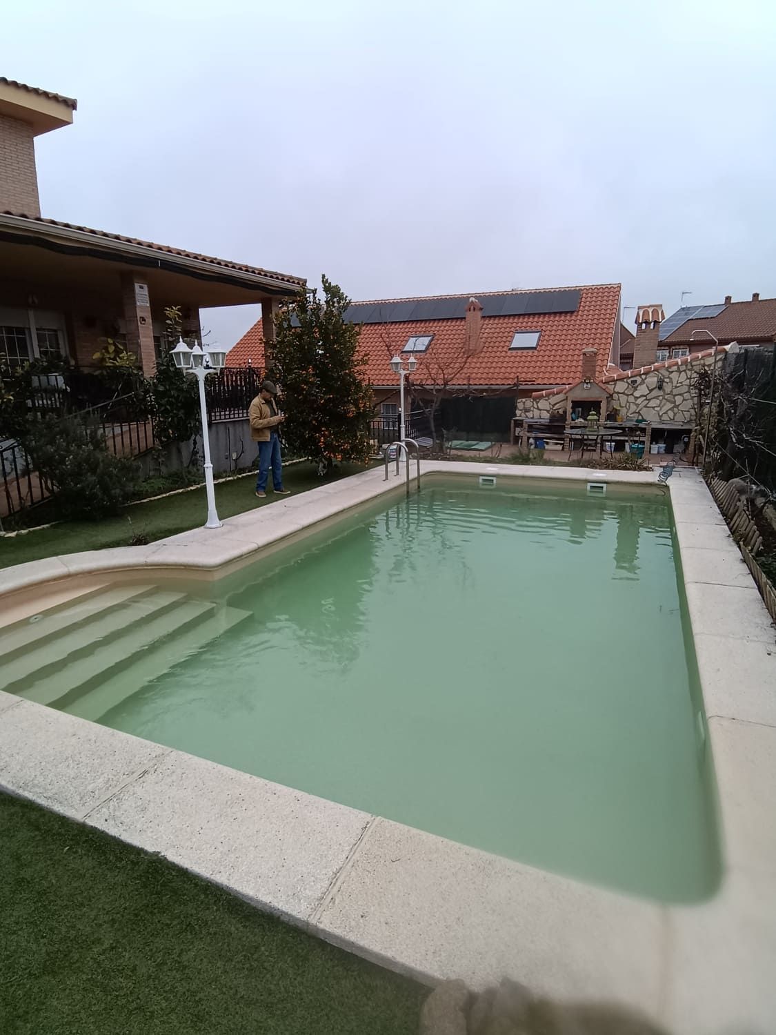 Piscina en un patio trasero con una casa, escalones y una persona cerca del borde; cielo nublado.