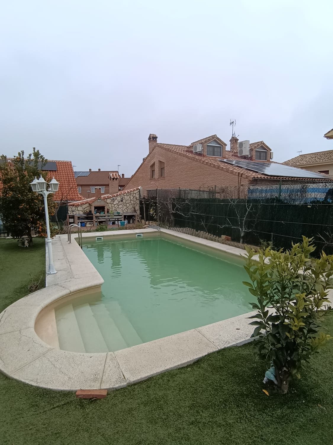 Piscina en un patio trasero con escaleras, rodeada de césped verde y edificios bajo un cielo nublado.