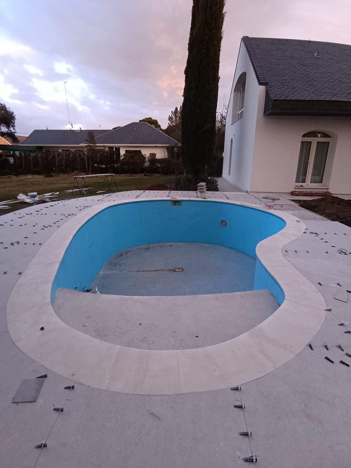 Piscina vacía, con forma de riñón, con interior azul claro, rodeada por un deck de hormigón gris, junto a una casa blanca.