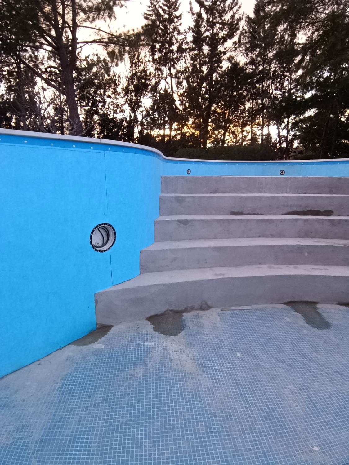 Interior de piscina azul con escaleras y tubería. Árboles al fondo.