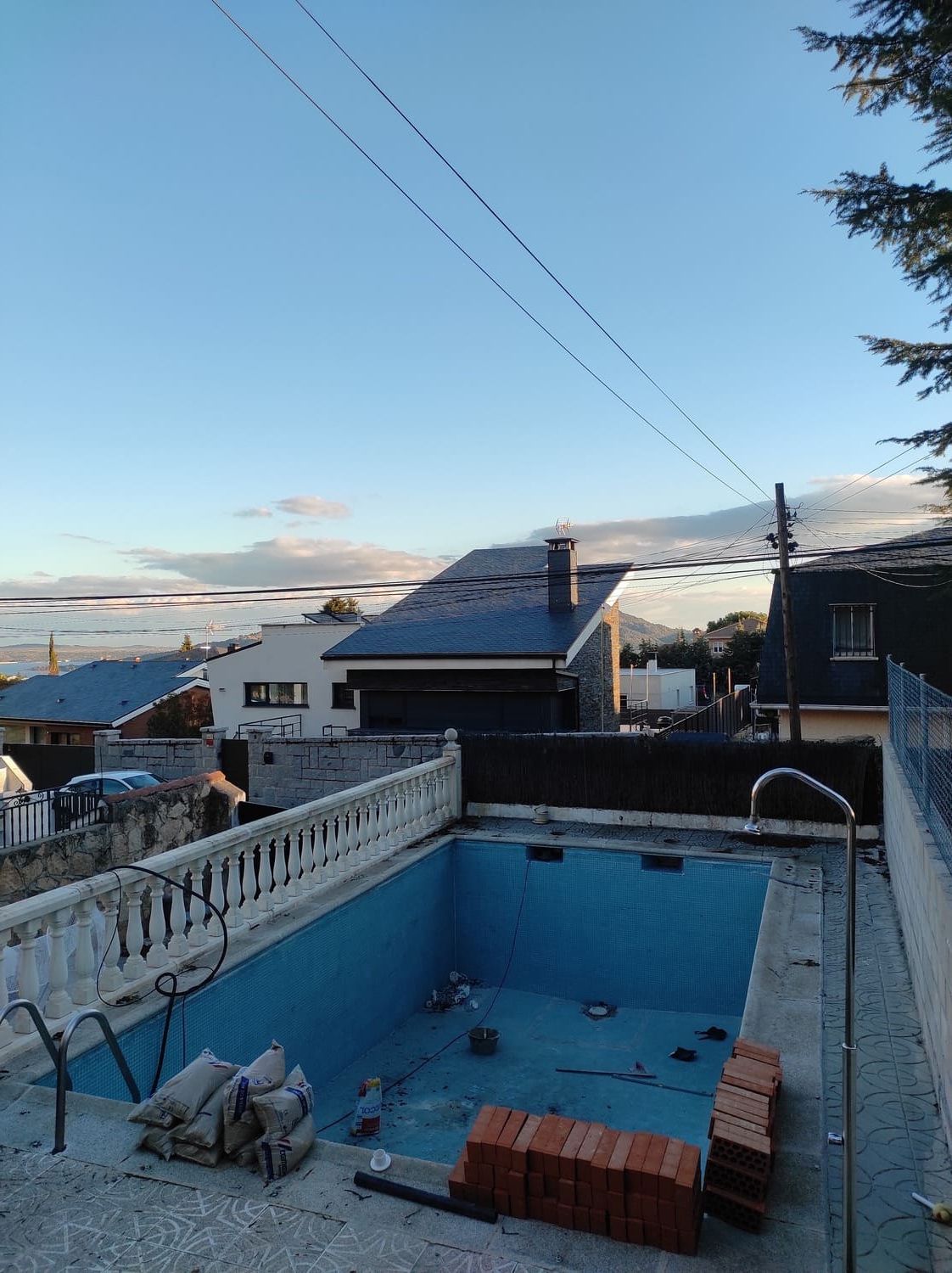 Piscina azul vacía con ladrillos, bolsas y edificios al fondo bajo un cielo nublado.