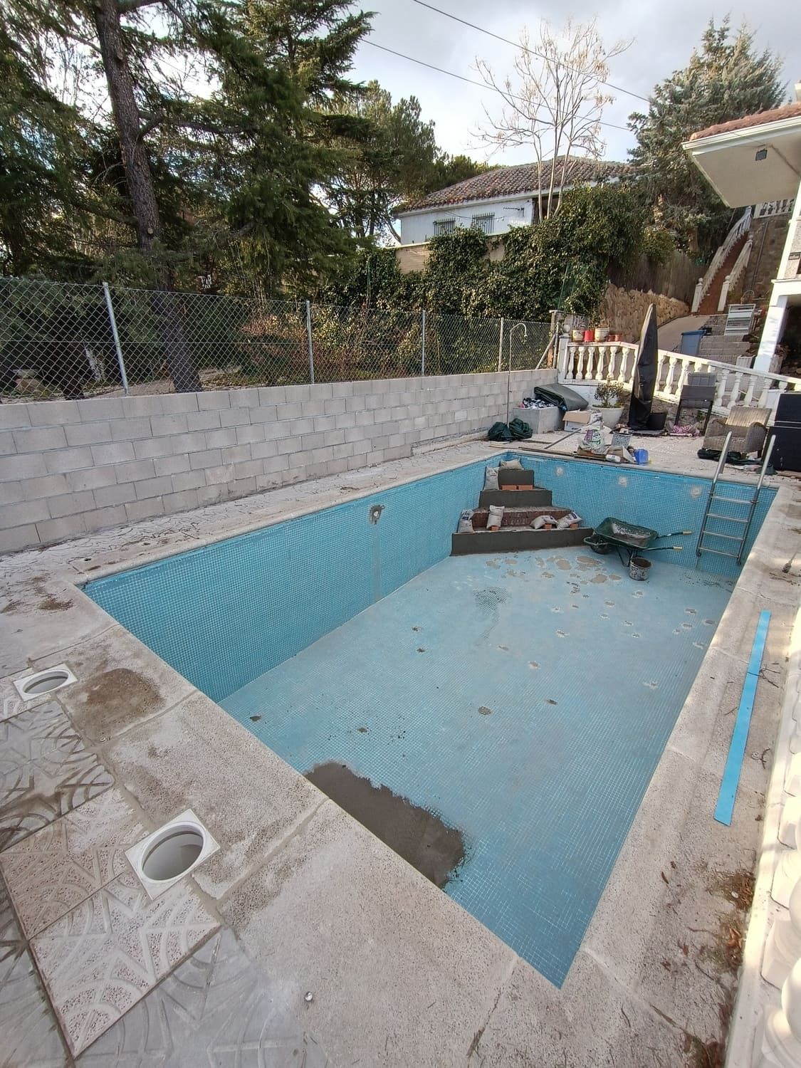 Piscina vacía con azulejos azules, escaleras y hormigón alrededor. Al fondo, una casa y árboles.