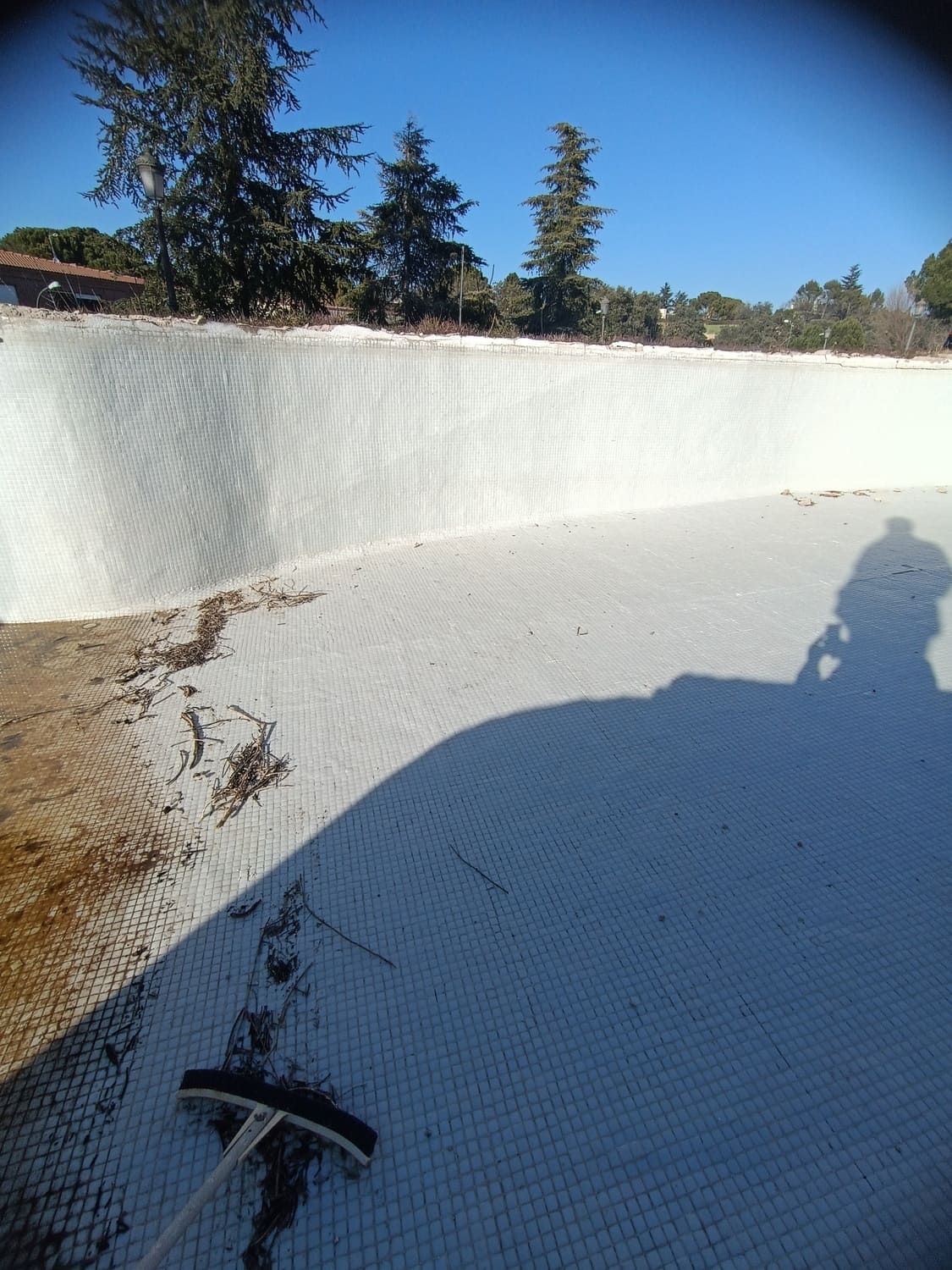 Piscina vacía con revestimiento blanco, limpiada con un cepillo. Árboles y cielo azul al fondo.
