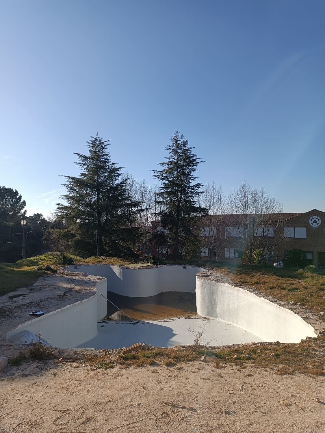 Piscina vacía, de paredes blancas y suelo de tierra, en una zona de césped con árboles, bajo un cielo azul.