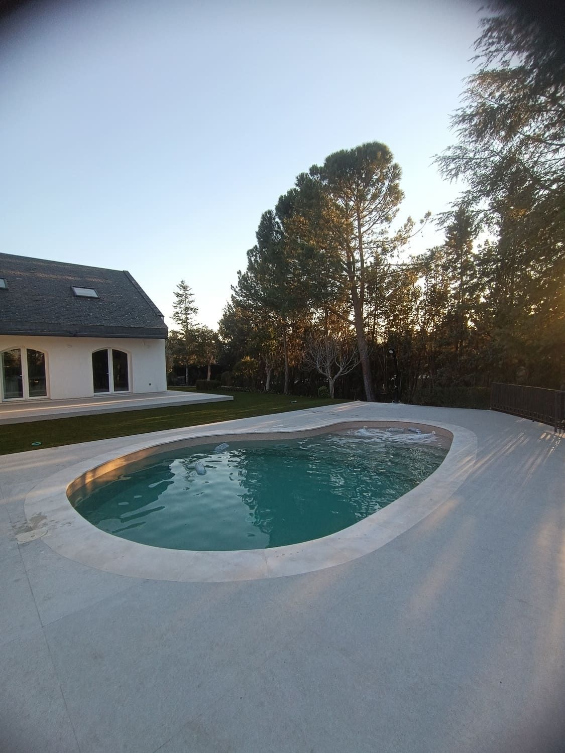 Piscina con marco de hormigón color claro, casa al fondo, árboles y agua azul.