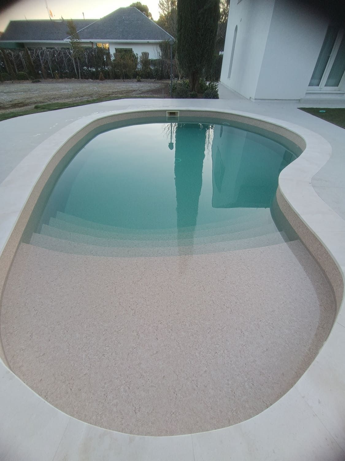 Piscina con superficie moteada de color claro y escalones, llena de agua cristalina. Está rodeada de hormigón blanco.