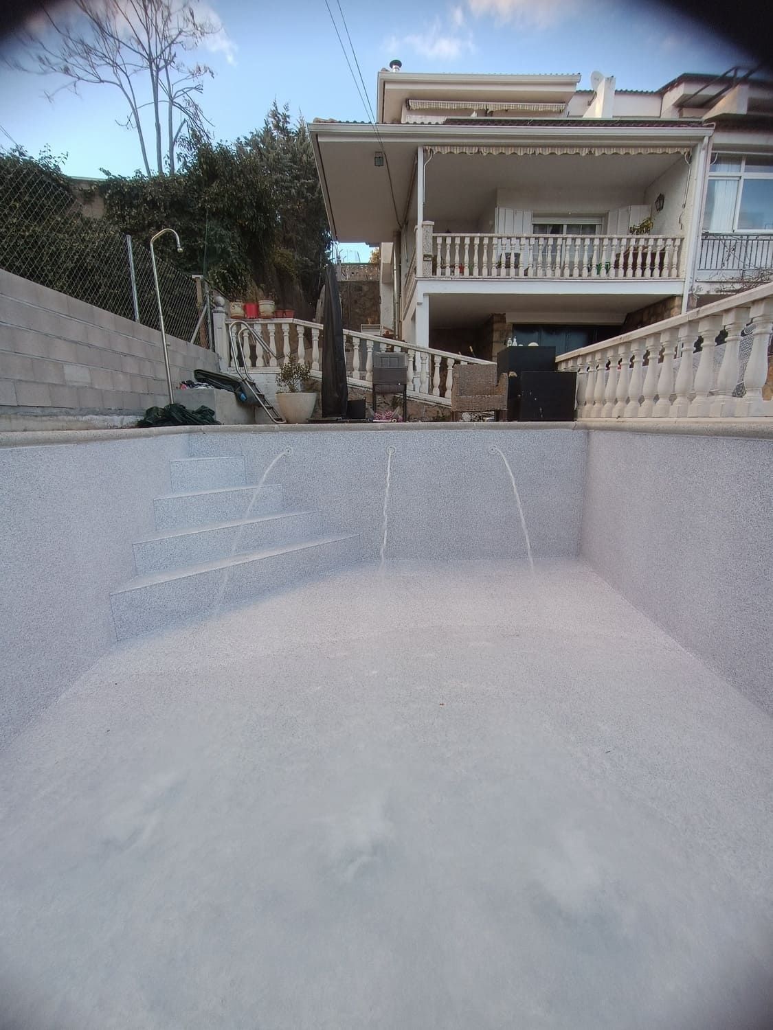 Piscina vacía con agua fluyendo, frente a una casa. Tonos blancos y grises, día soleado.