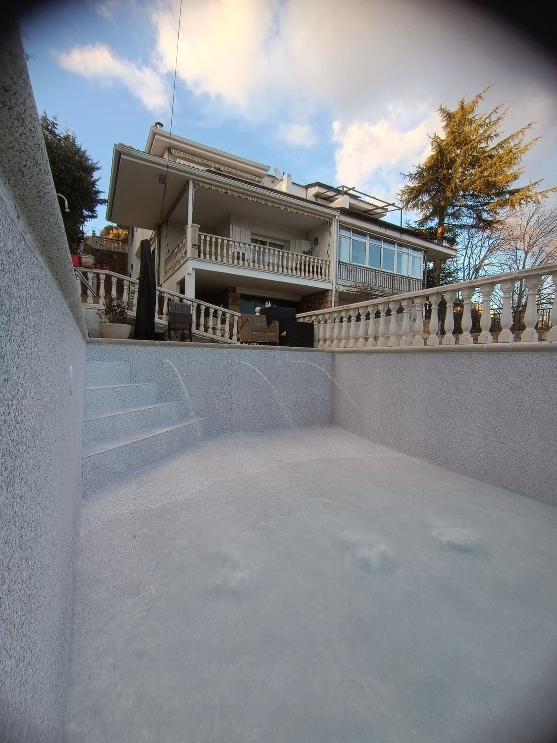 Piscina vacía frente a una casa blanca de varios pisos con un balcón bajo un cielo azul.