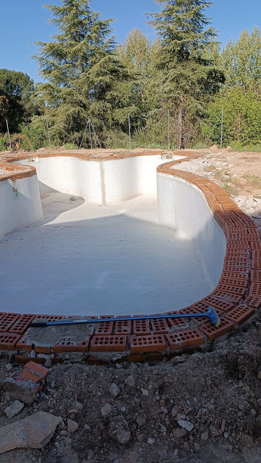 Construcción de piscina de canto curvo, base de hormigón y paredes blancas, con borde de ladrillos rojos.