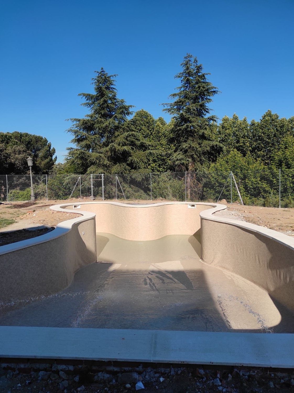 Piscina vacía, de nueva construcción, con paredes beige, bordes de hormigón y árboles de fondo.