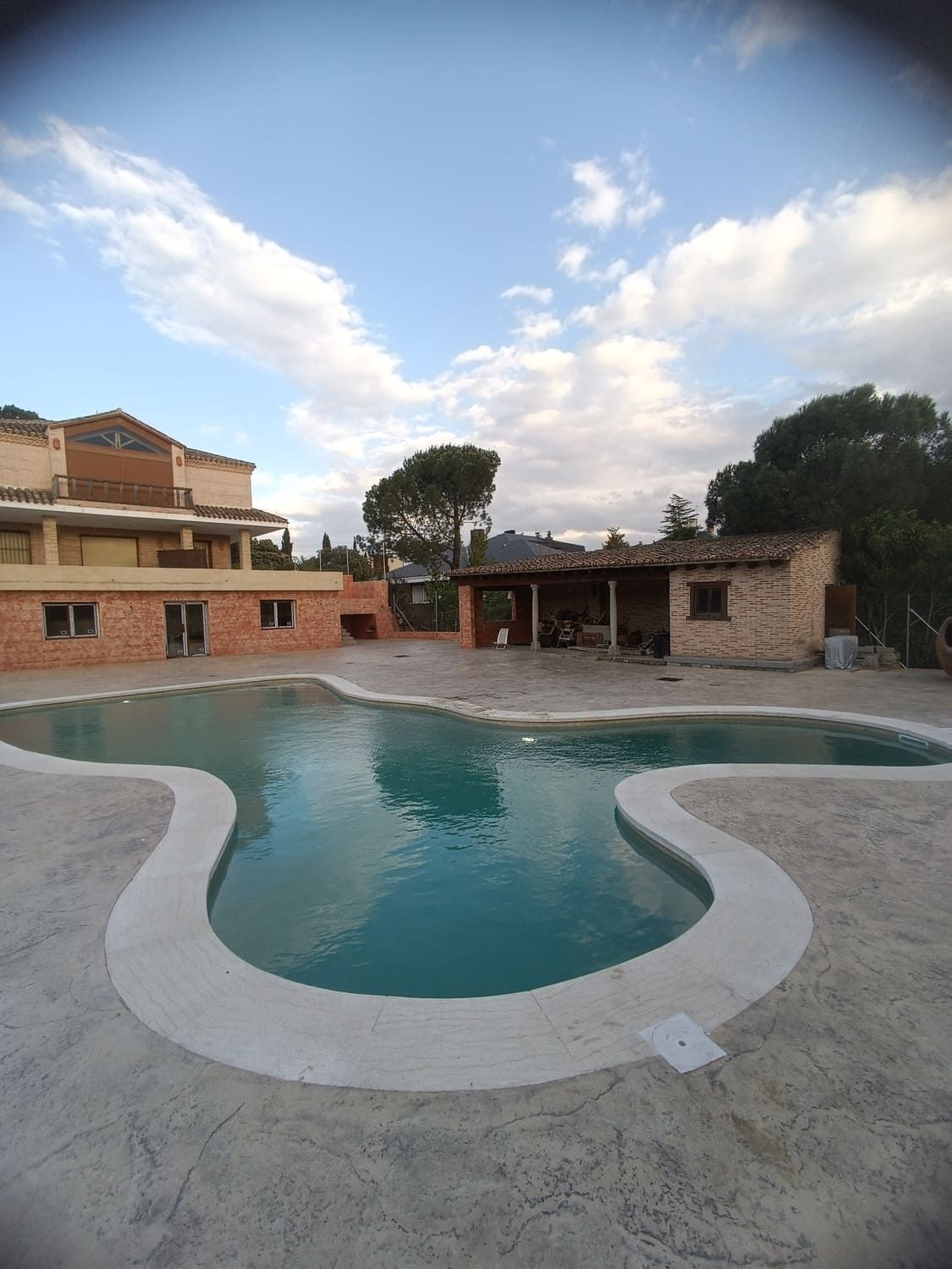 Piscina con bordes curvos en un patio de concreto, con edificios y árboles bajo un cielo nublado.