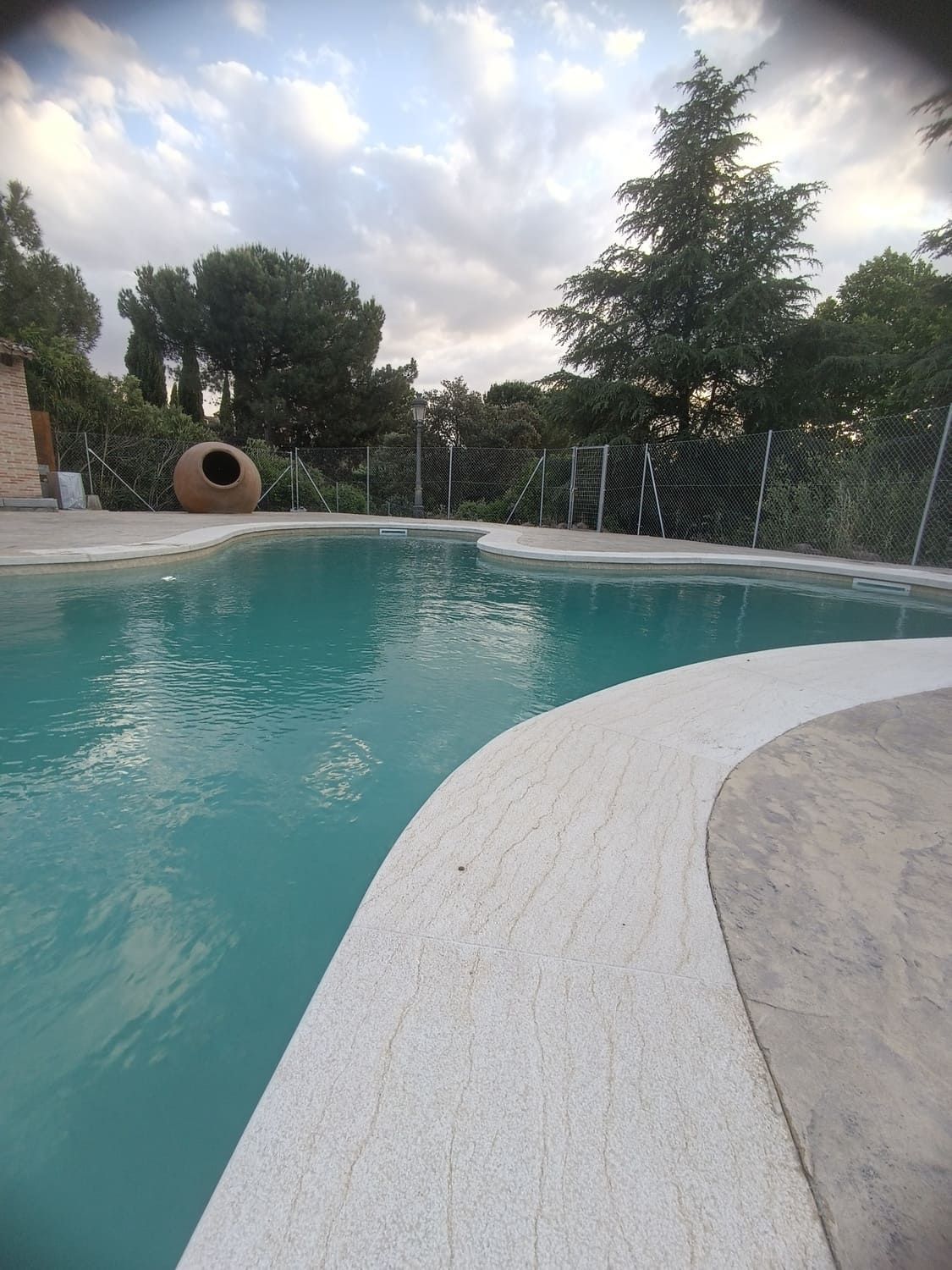 Piscina con agua azul verdosa, borde de piedra y cielo nublado en un entorno natural.
