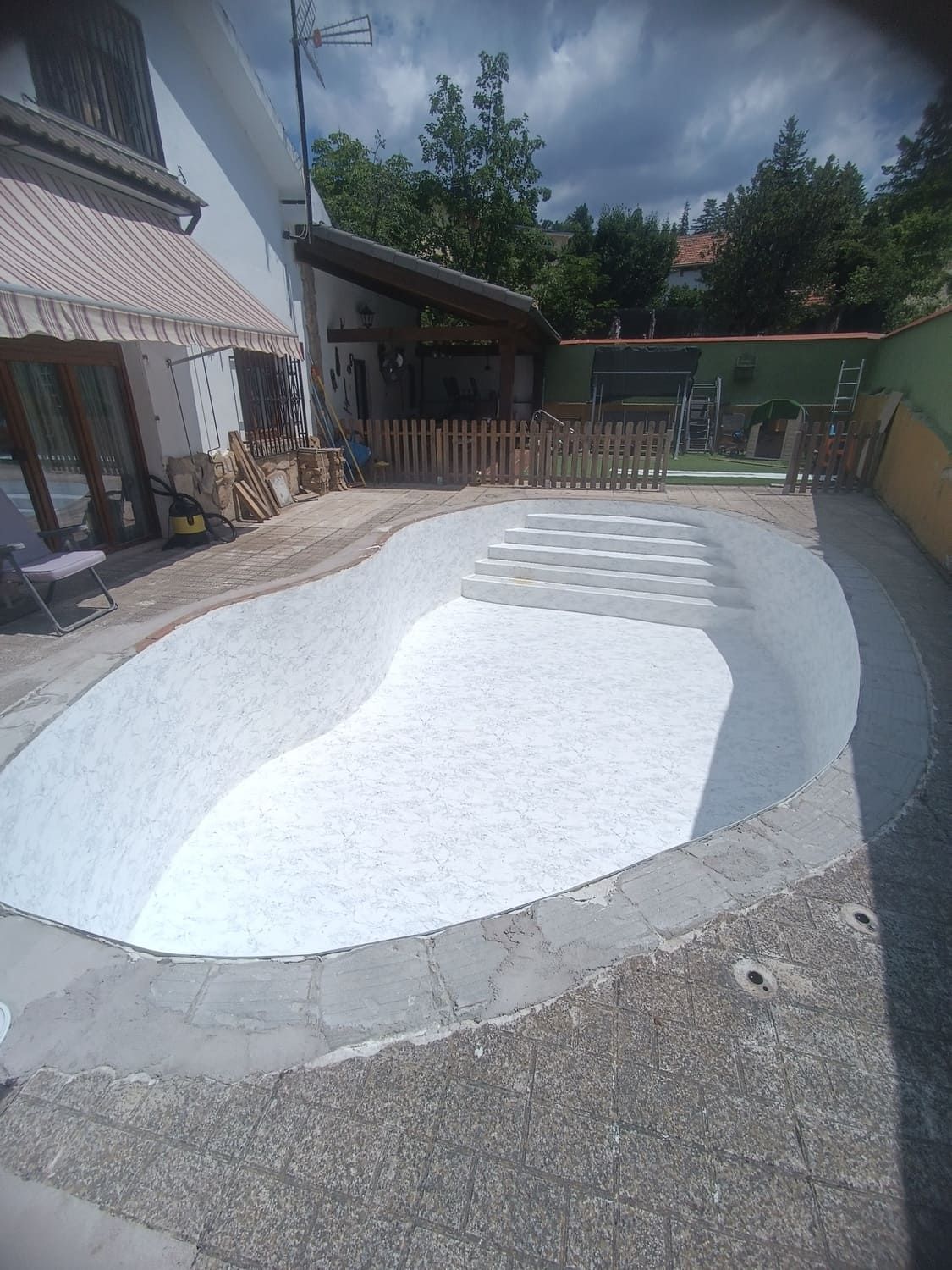 Piscina vacía, pintada de blanco, con forma de riñón, con escalones, rodeada de un borde de hormigón y una pequeña valla de madera.