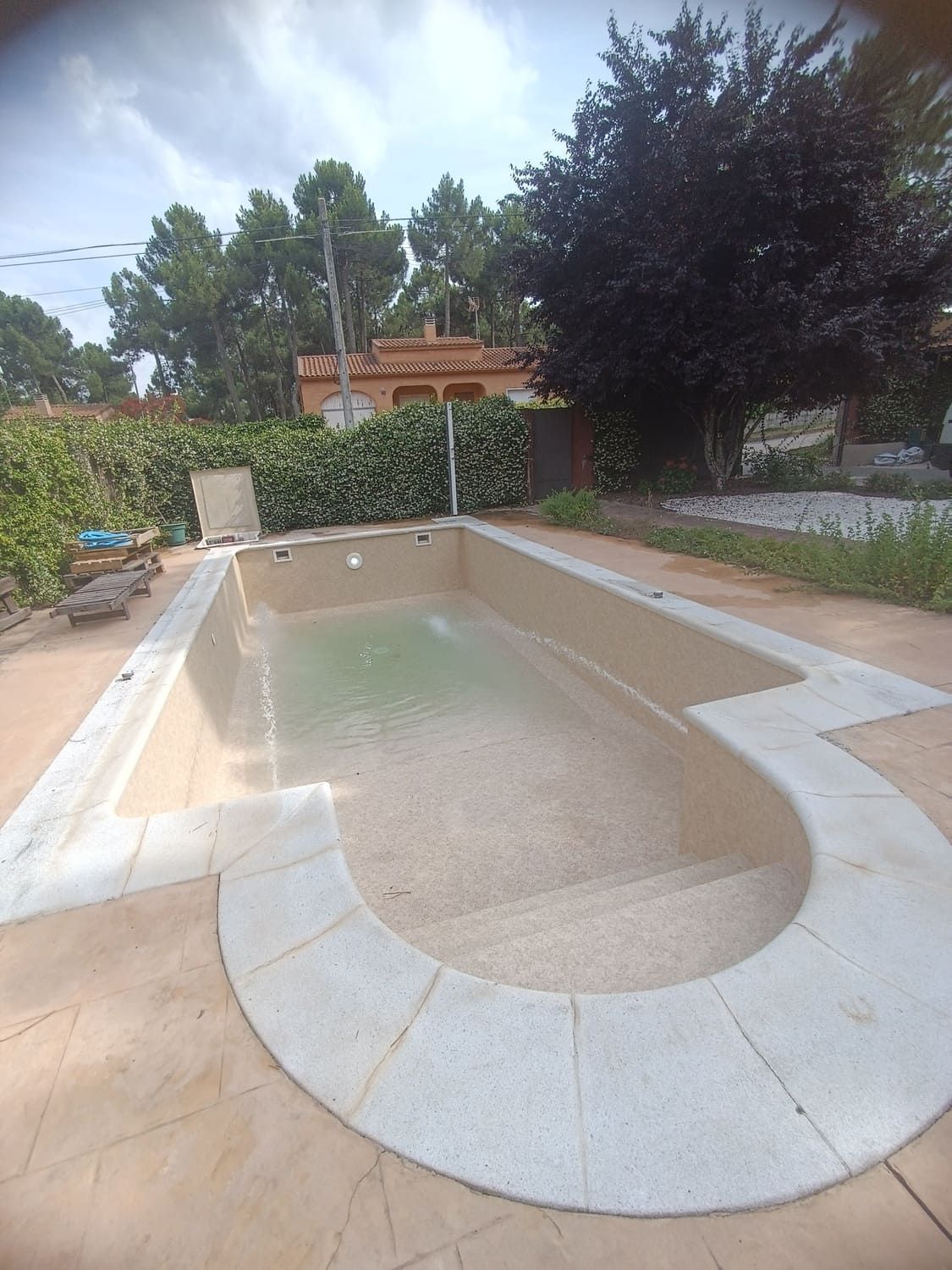 Piscina vacía rectangular con escalones curvos, rodeada de piedra, con árboles y un edificio al fondo.