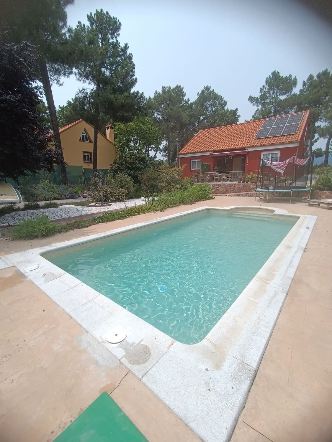 Piscina rectangular frente a una casa roja con paneles solares. Árboles y otro edificio amarillo al fondo.