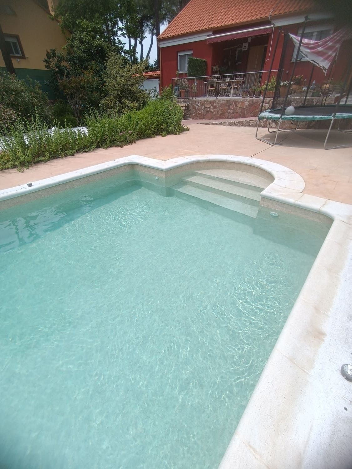 Piscina con escaleras, rodeada de un patio y una casa roja.