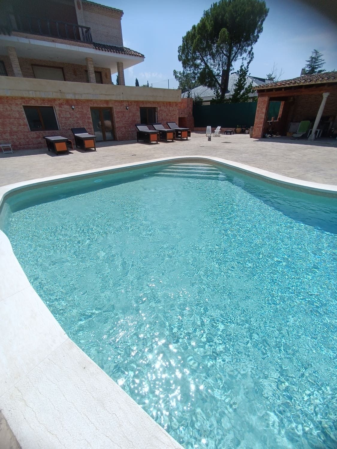 Una piscina rectangular de agua turquesa cristalina. Sillas de descanso en el borde. Un edificio de ladrillo al fondo. Día soleado.