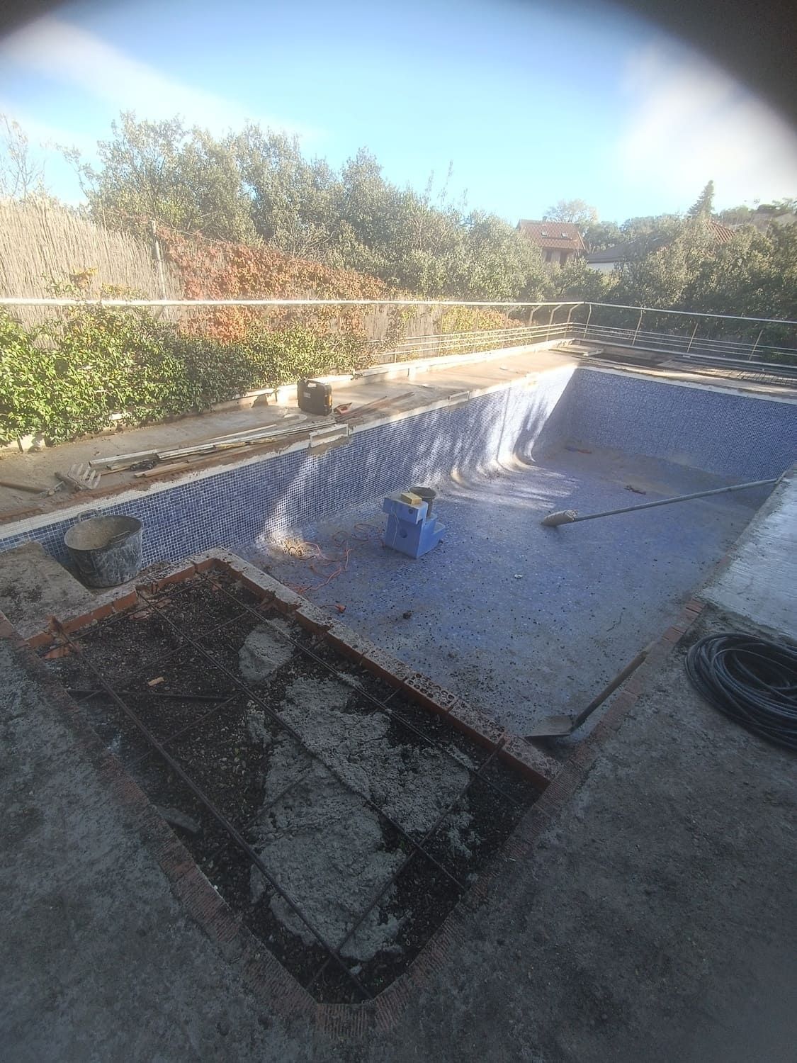 Una piscina vacía y embaldosada, rodeada de hormigón y vegetación crecida bajo un cielo soleado.