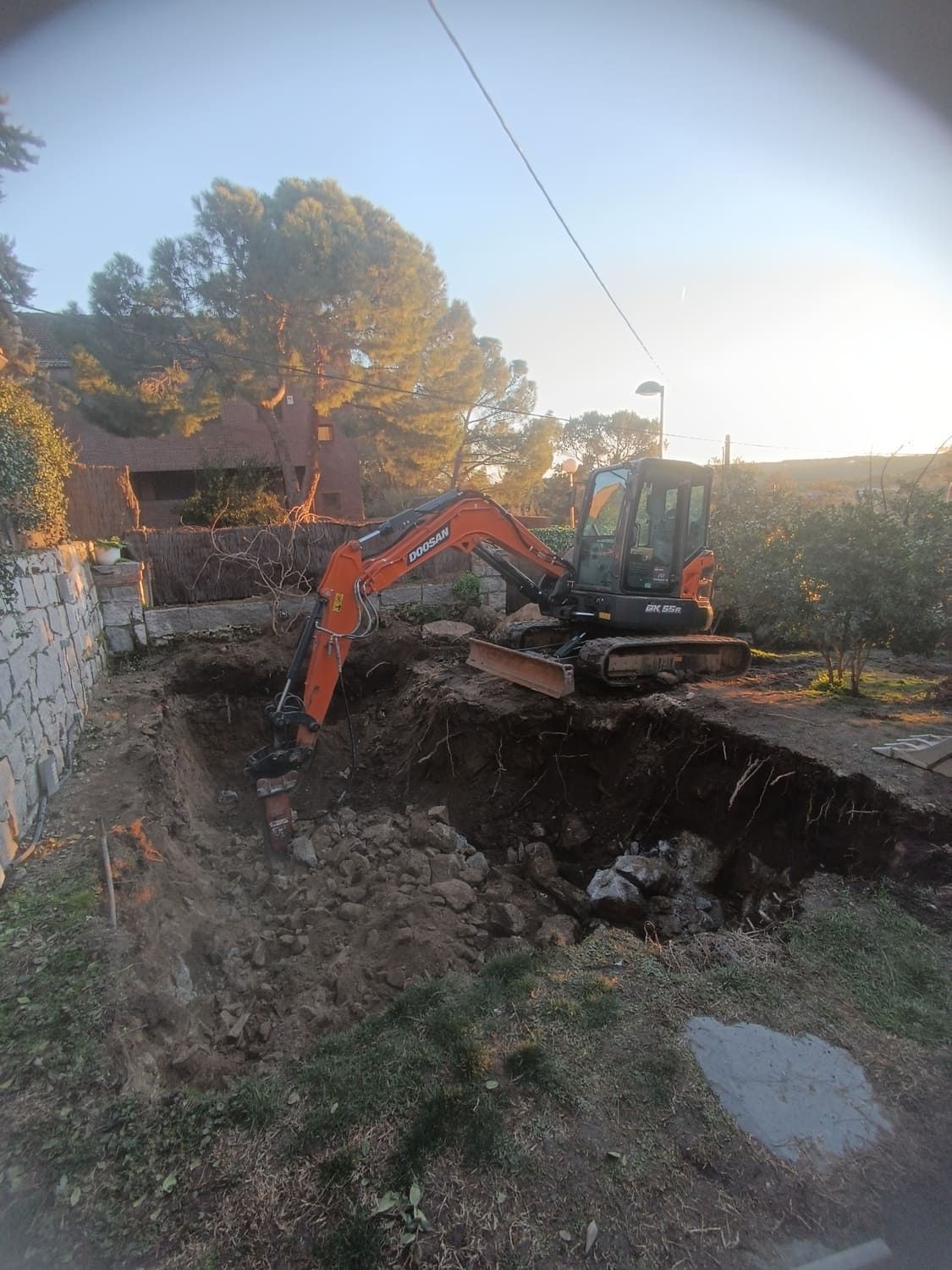 Una excavadora excavando en un pozo de tierra al aire libre. Brazo naranja, máquina gris y negra. Luz solar brillante, follaje y una casa al fondo.
